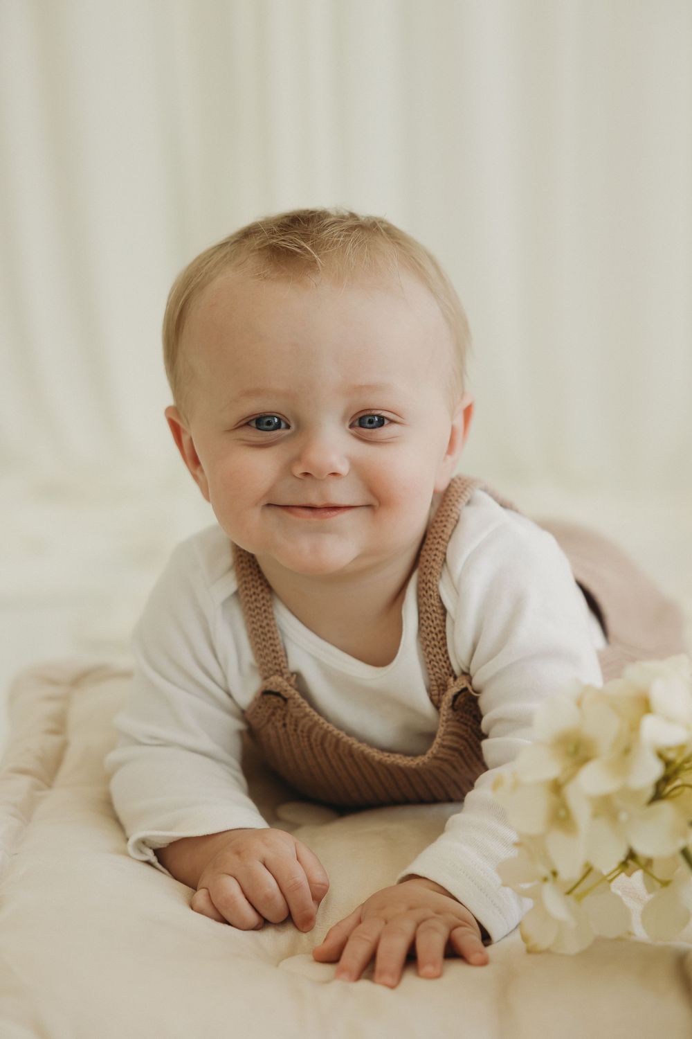 Smiling baby in white onesie and brown overalls lying on a soft blanket with white flowers nearby.