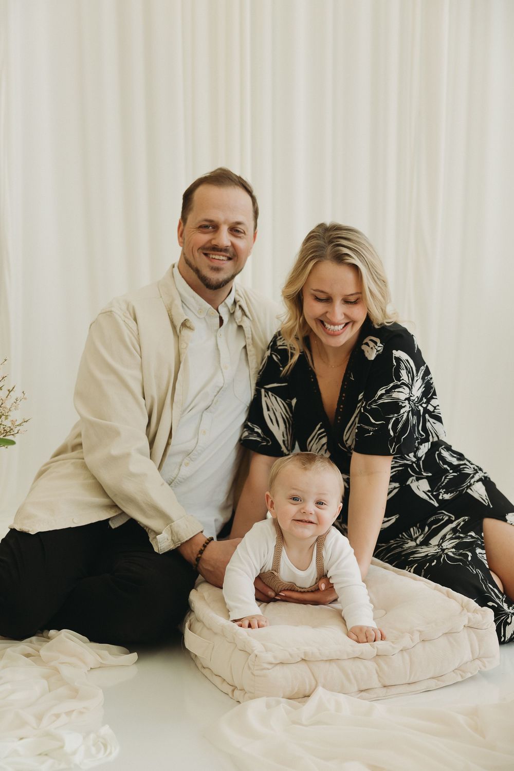 Happy family portrait with parents and baby sitting on a cushion against a white curtain backdrop.