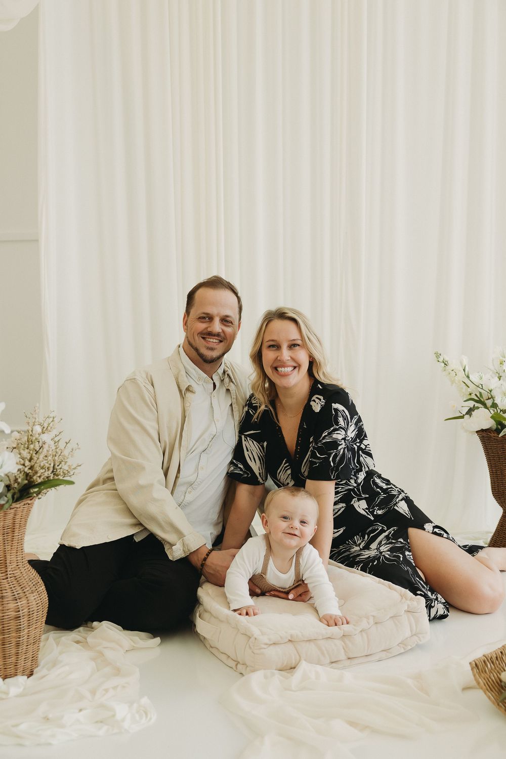 A smiling family with a man, woman, and baby posing together on a white floor with floral backdrop.