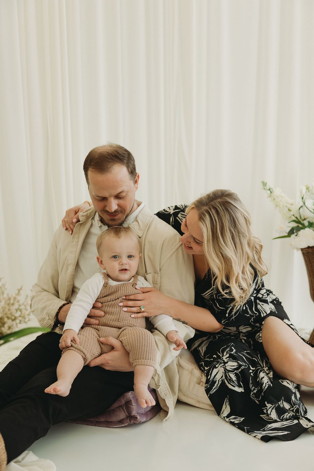 A couple sitting on the floor with their baby, man in beige suit, woman in floral dress, white curtain backdrop.