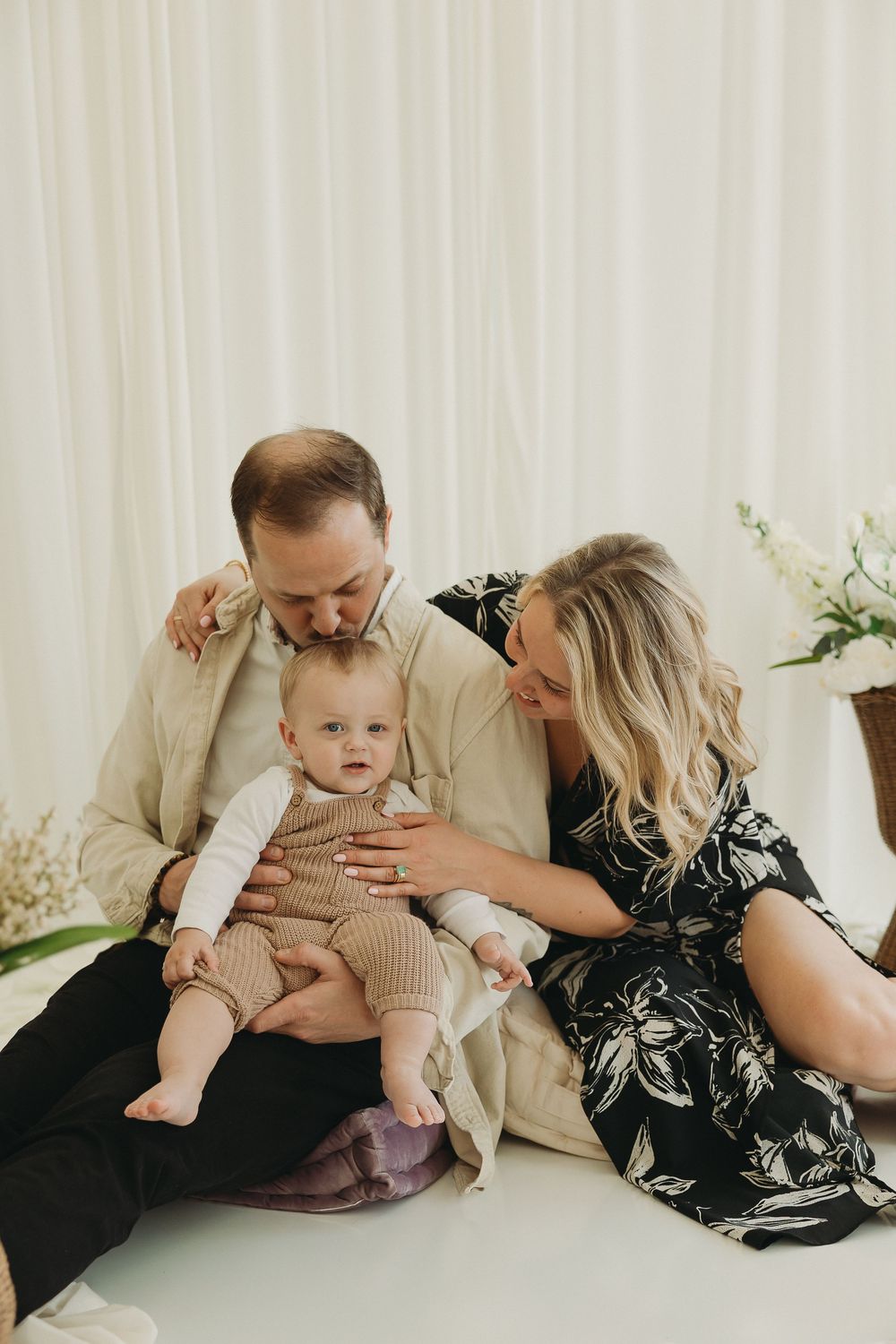 A couple sits on a white floor, lovingly gazing at their baby in a bright studio setting.