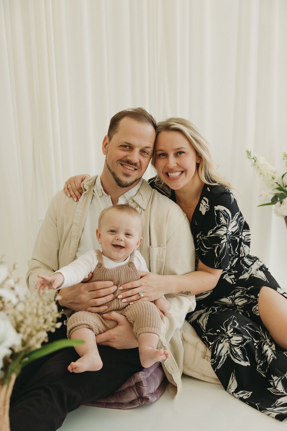 Happy family portrait with parents smiling and baby laughing, seated against white curtain backdrop in studio setting.