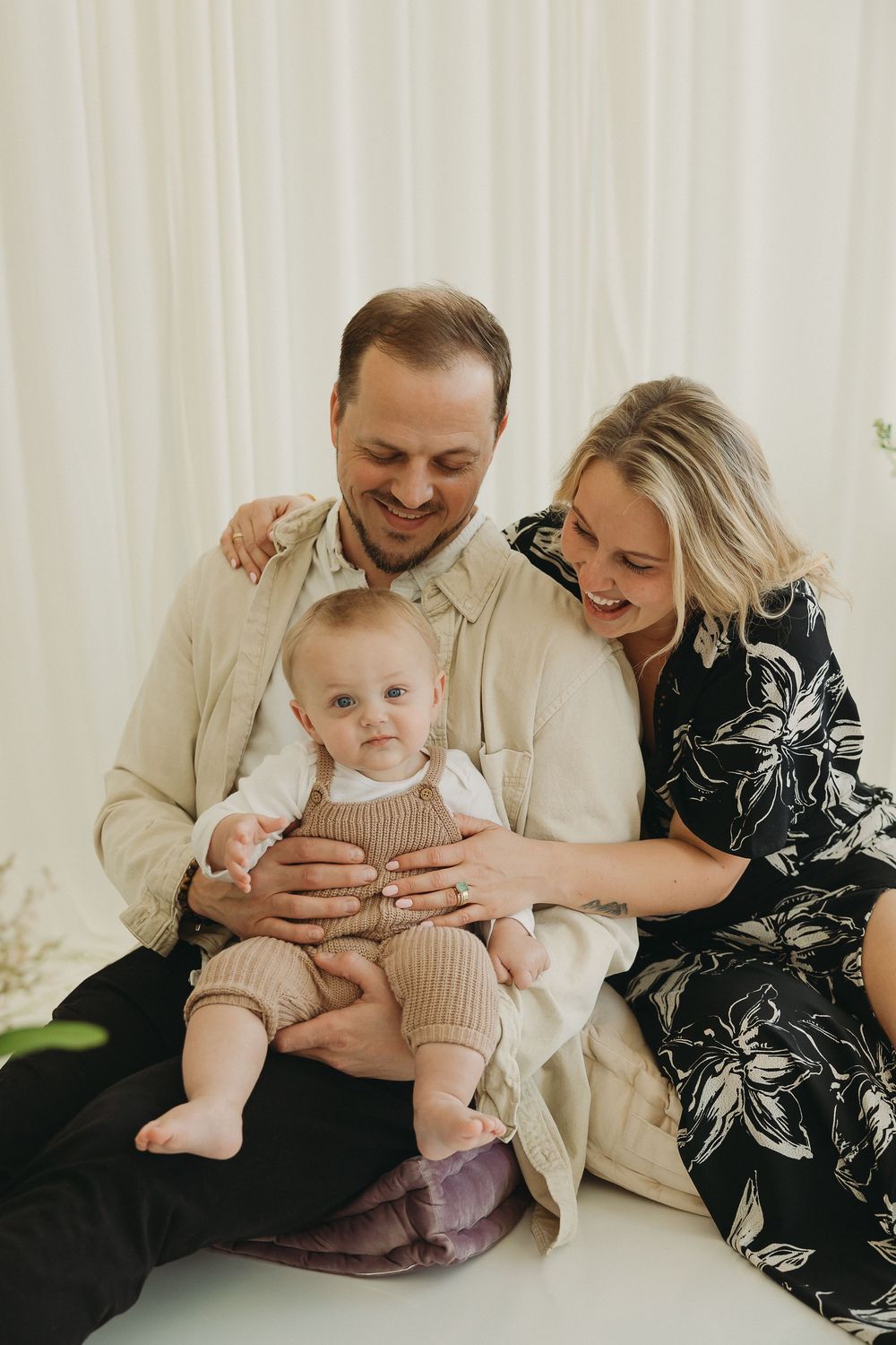 A smiling family with a baby in a beige romper sitting on dad's lap, mom in floral dress, against white curtains.