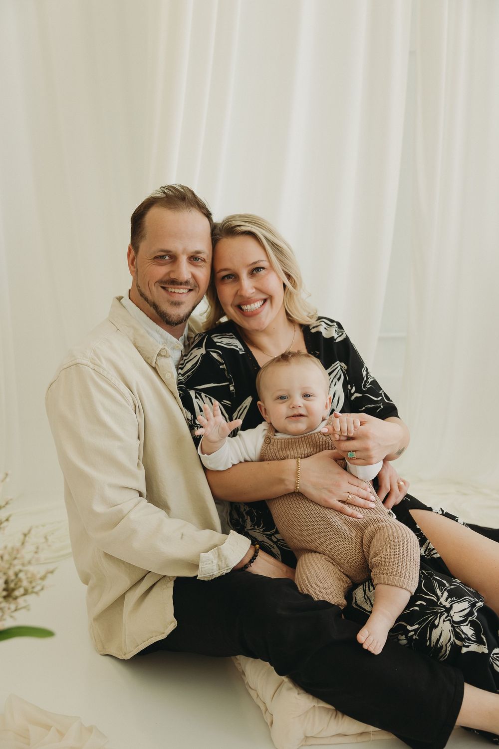 A smiling couple sits together on the floor holding their happy baby in a bright white studio portrait setting.