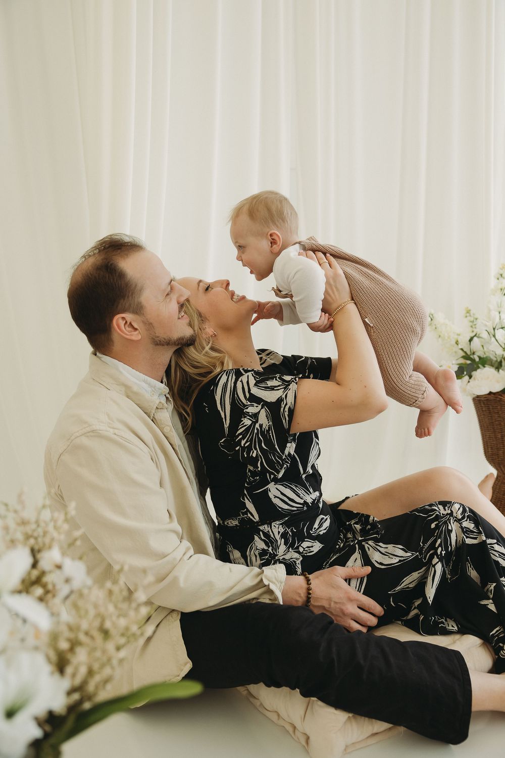 A couple sits on the floor holding their baby up, sharing a joyful family moment in a bright, airy room.