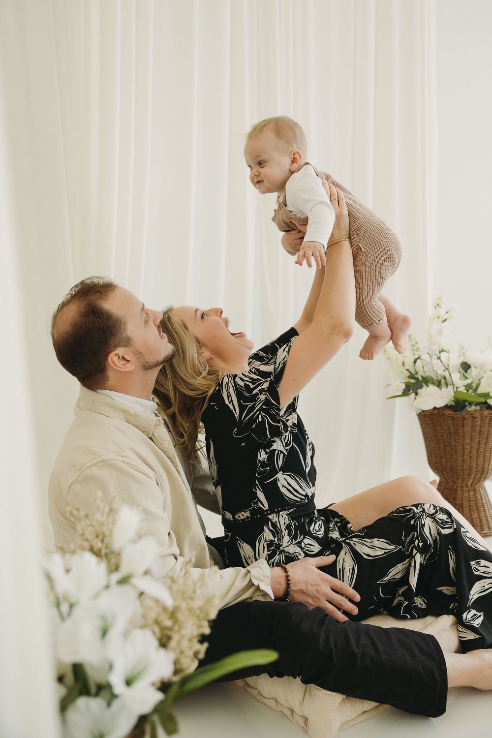 A couple sits on the floor lifting their smiling baby in the air, surrounded by white flowers and curtains.