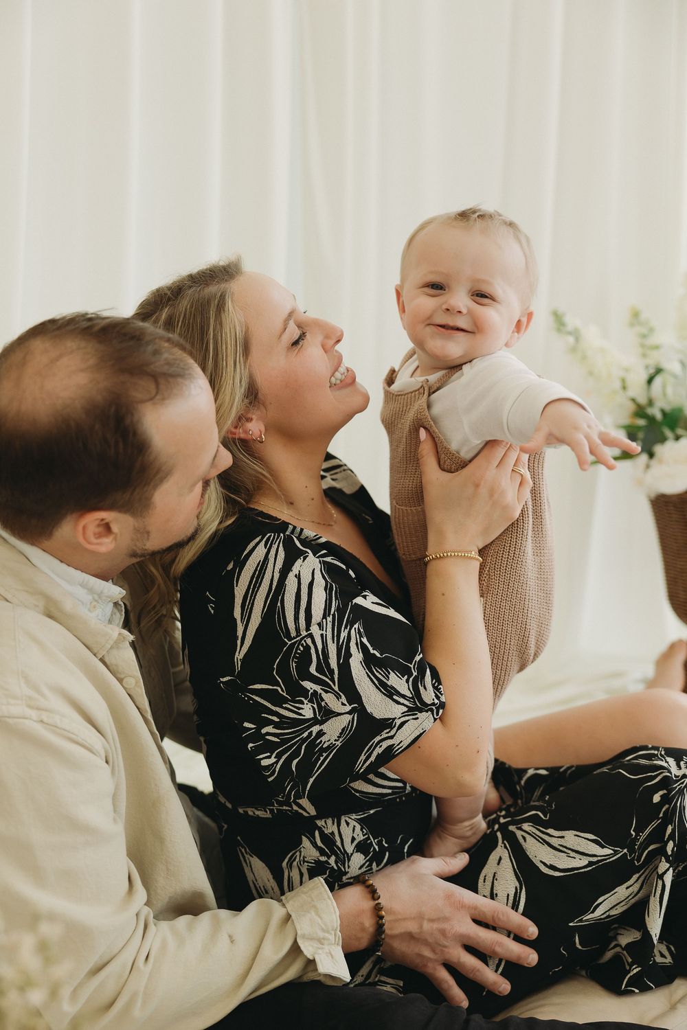 A happy family with a smiling baby being held up by the mother while the father looks on lovingly.