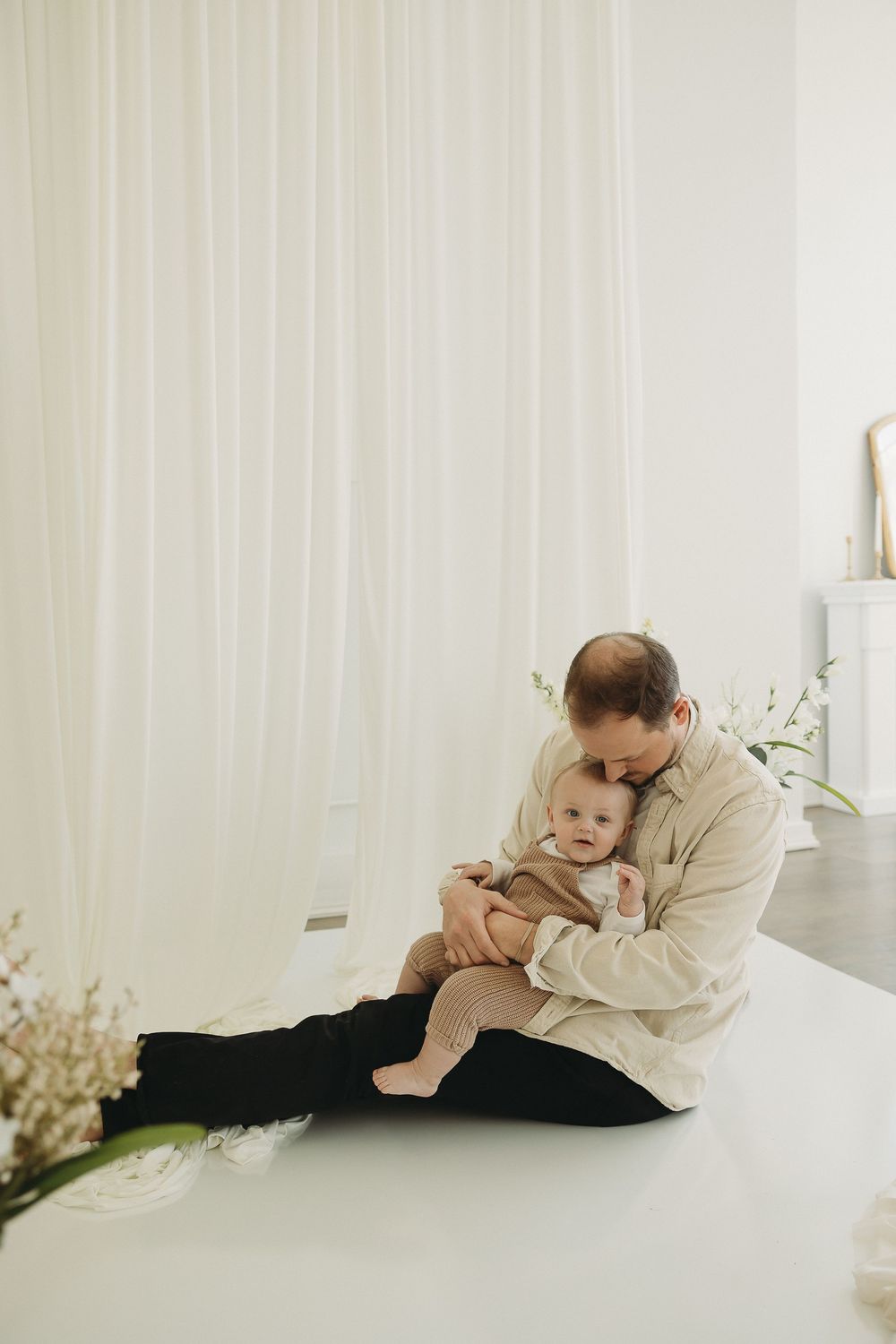 A father sits on a white floor cradling his baby, surrounded by soft white curtains and flowers.