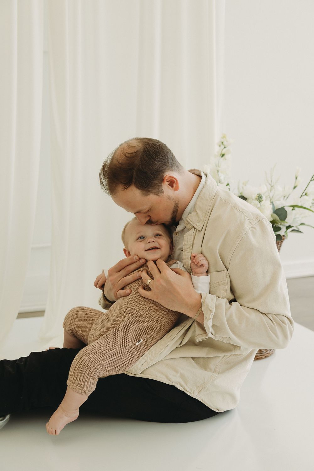 A father sits on the floor kissing his smiling baby, both dressed in neutral tones in a bright white room.