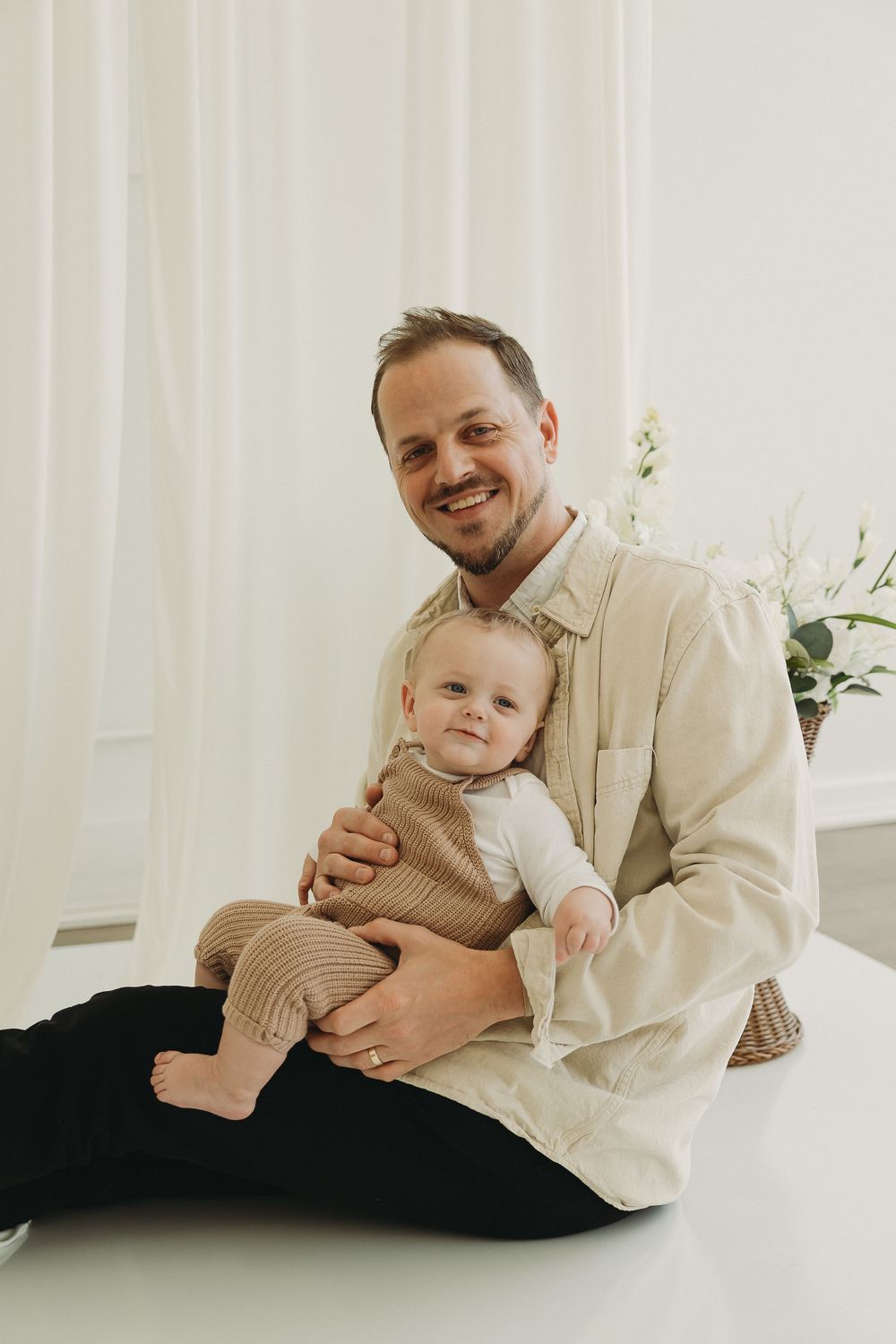 Smiling father sitting on floor holding happy baby in brown outfit against white curtain backdrop.