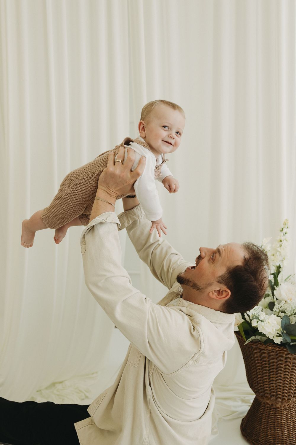 A joyful father lifts his smiling baby in the air in a bright, white room with flowers nearby.