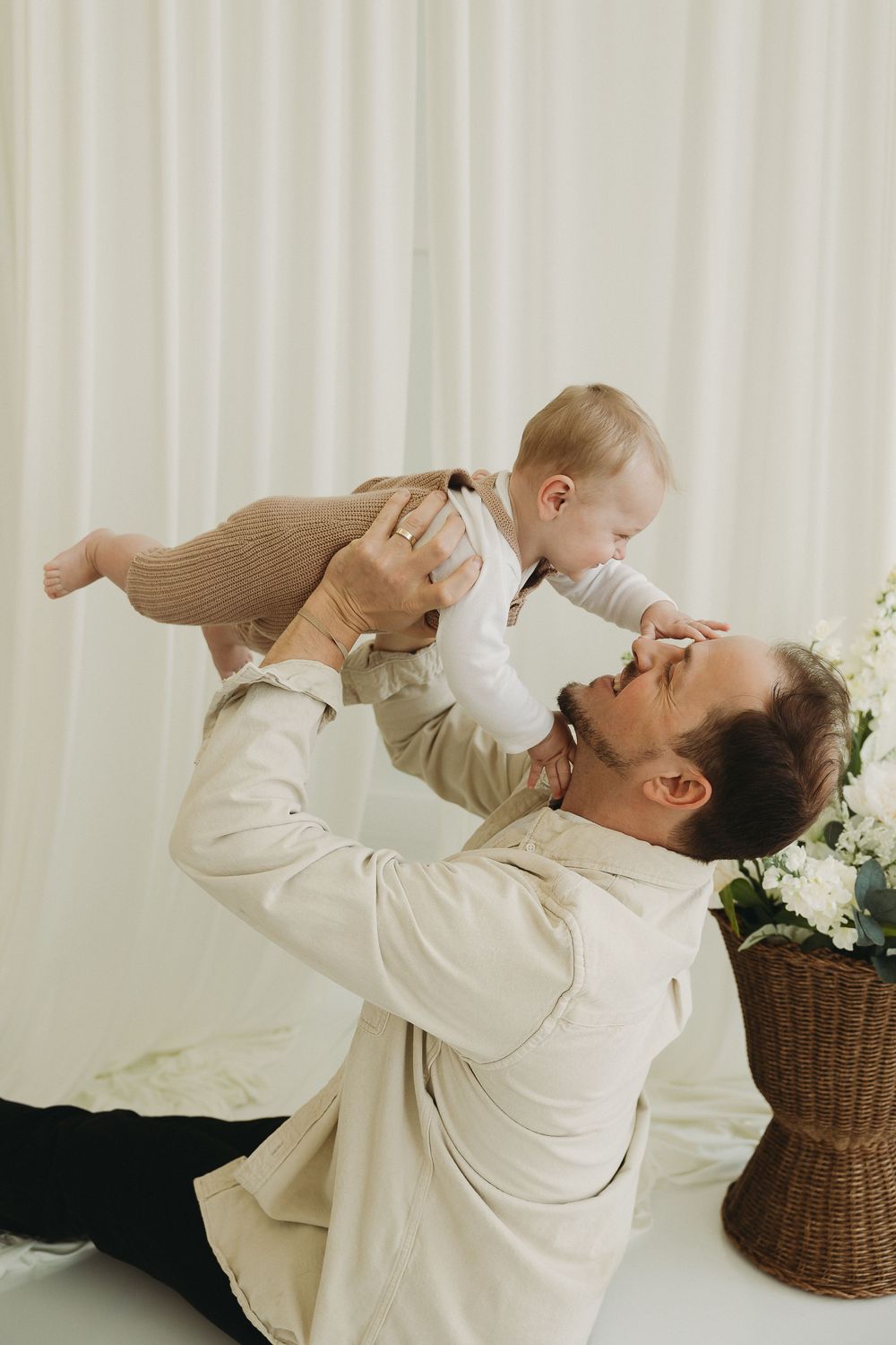 A father joyfully lifts his smiling baby in a bright, white studio setting with flowers nearby.