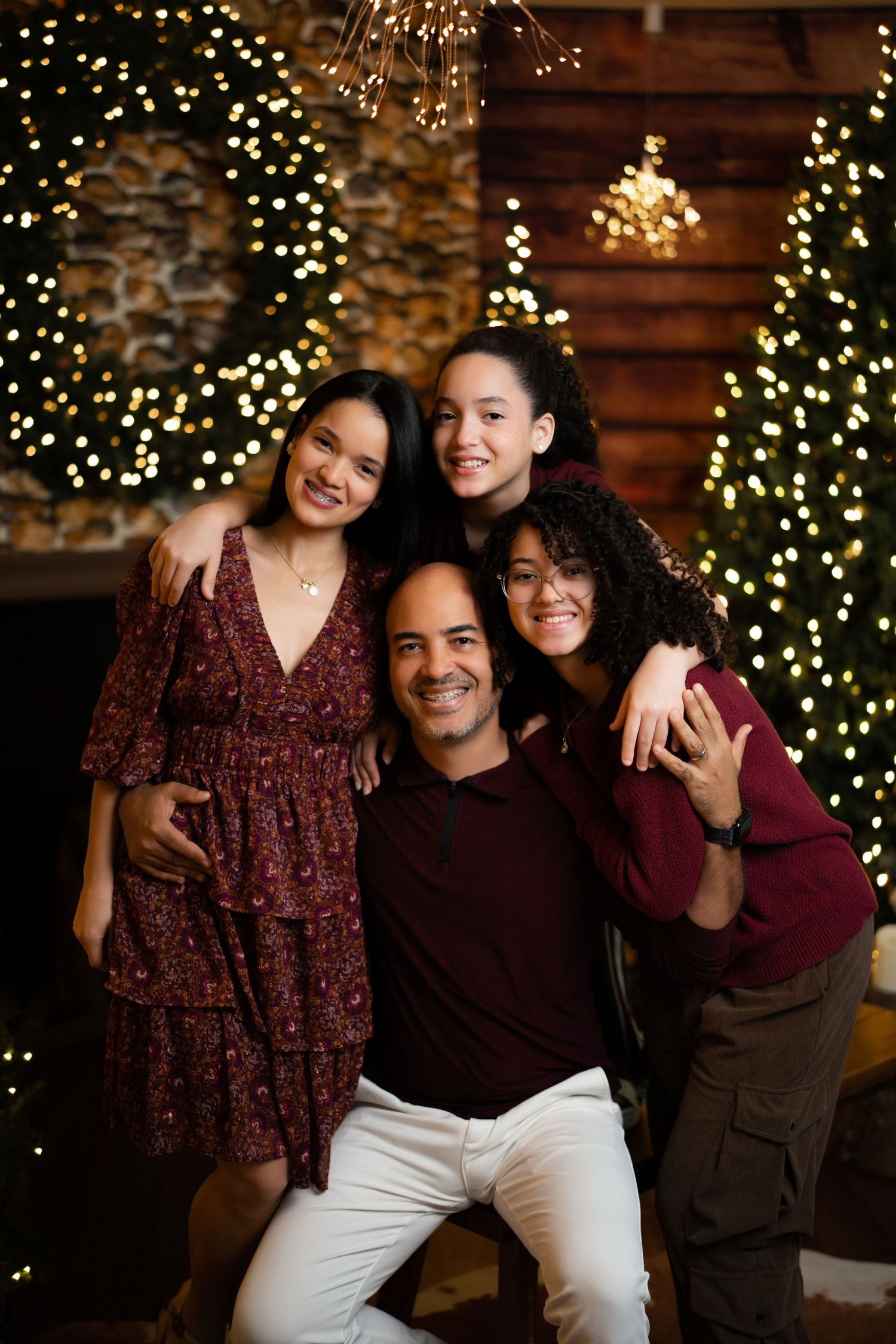 Family of four smiling together in front of decorated Christmas trees with warm bokeh lights.