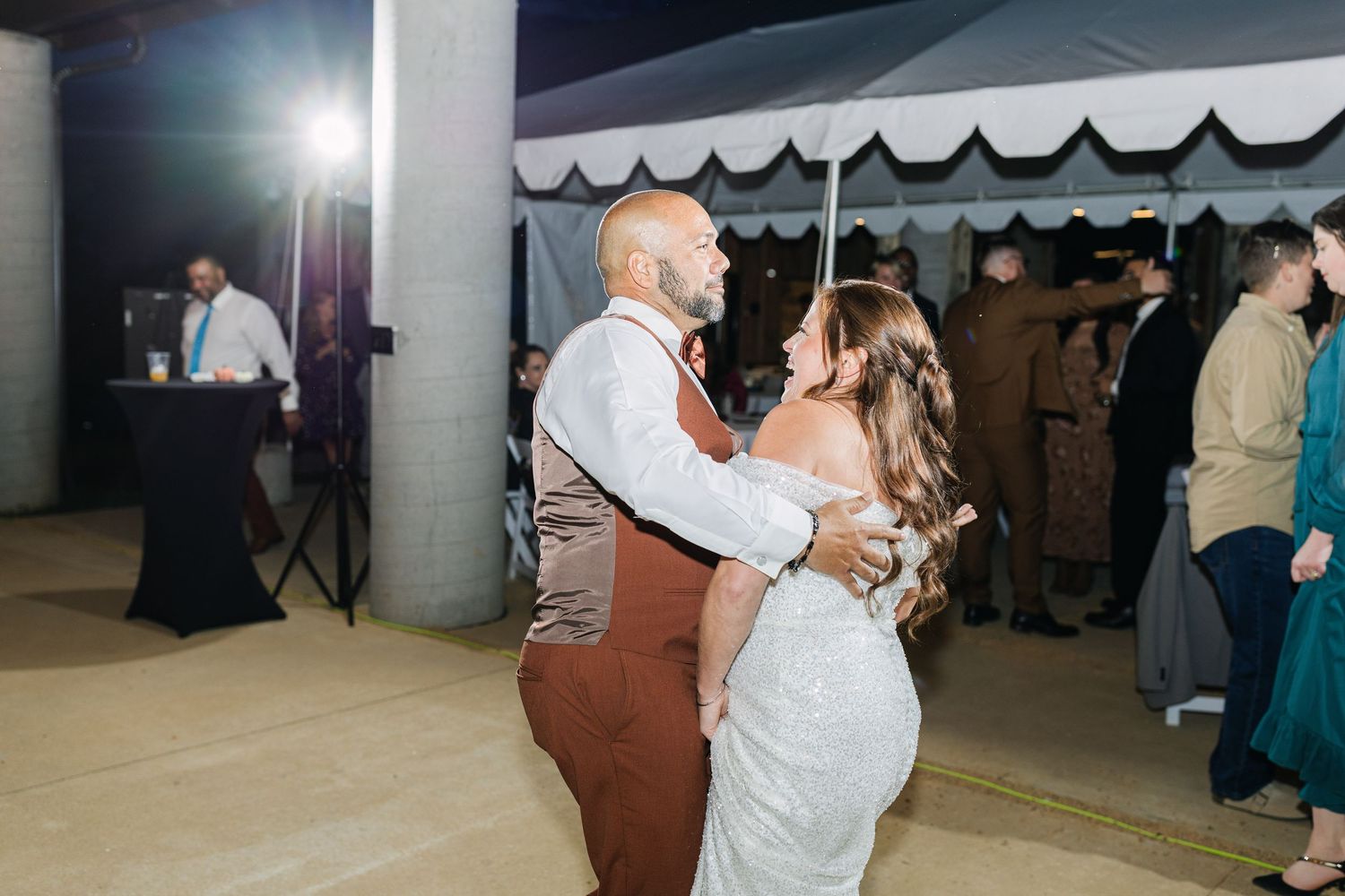 Bride and groom share their first dance at an outdoor wedding reception under a white tent at night.