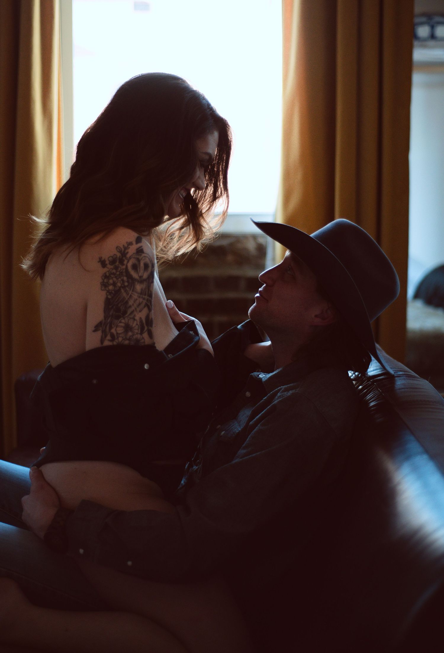 Intimate couple moment on sofa, tattooed woman leaning over man in cowboy hat near window with warm curtains.