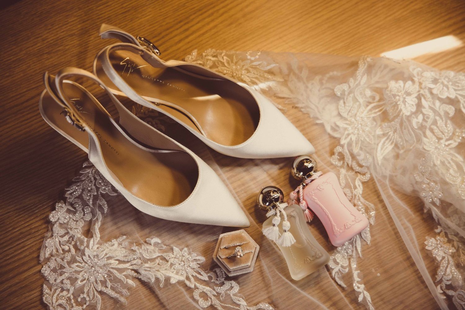Elegant white pointed-toe slingback wedding heels displayed with perfume, a ring box, and lace bridal veil on wood floor.