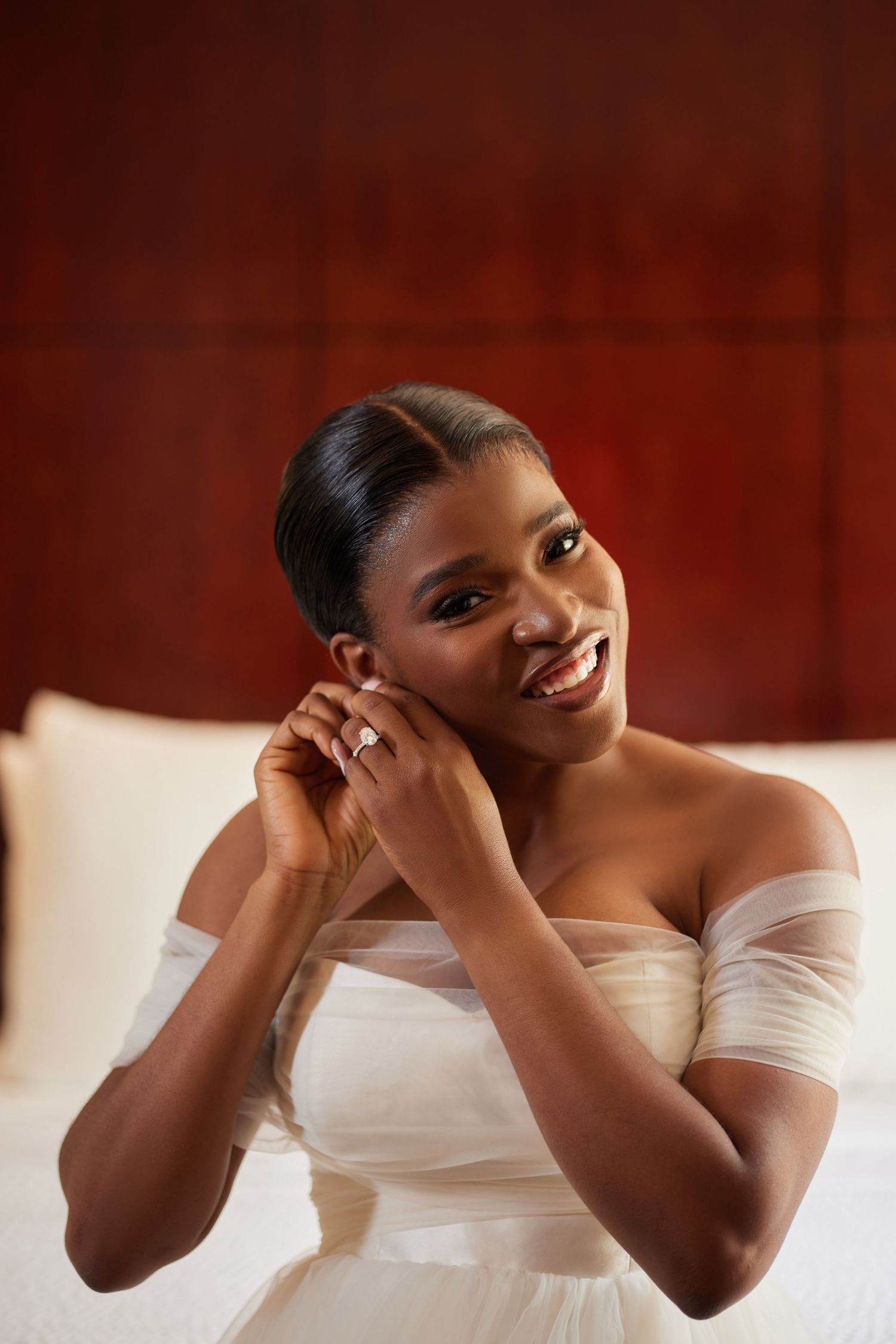Smiling Black bride in off-shoulder white gown putting on earrings while sitting on bed in elegant room.