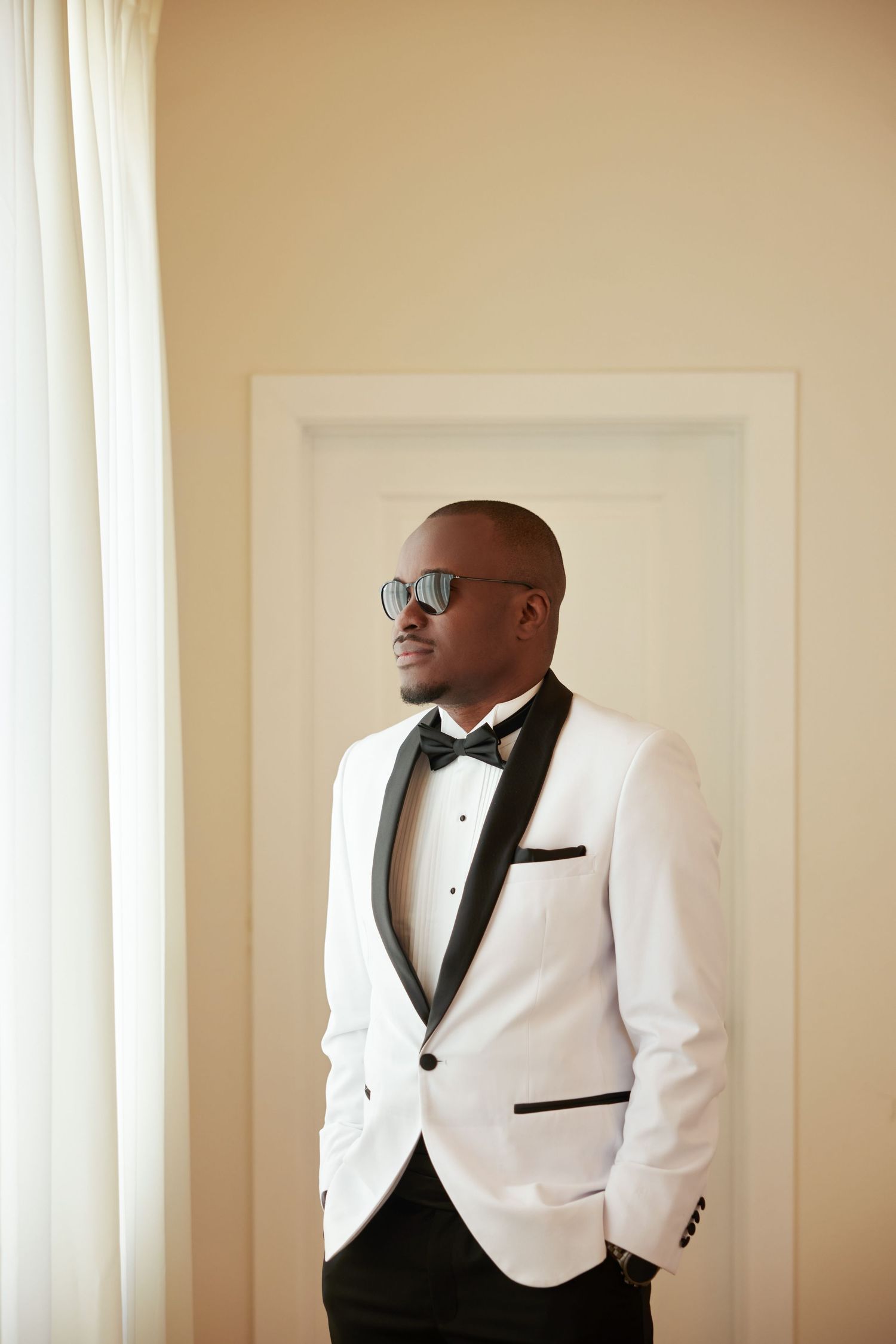 Stylish man in white tuxedo jacket with black lapels and bow tie, wearing sunglasses, standing near a window.