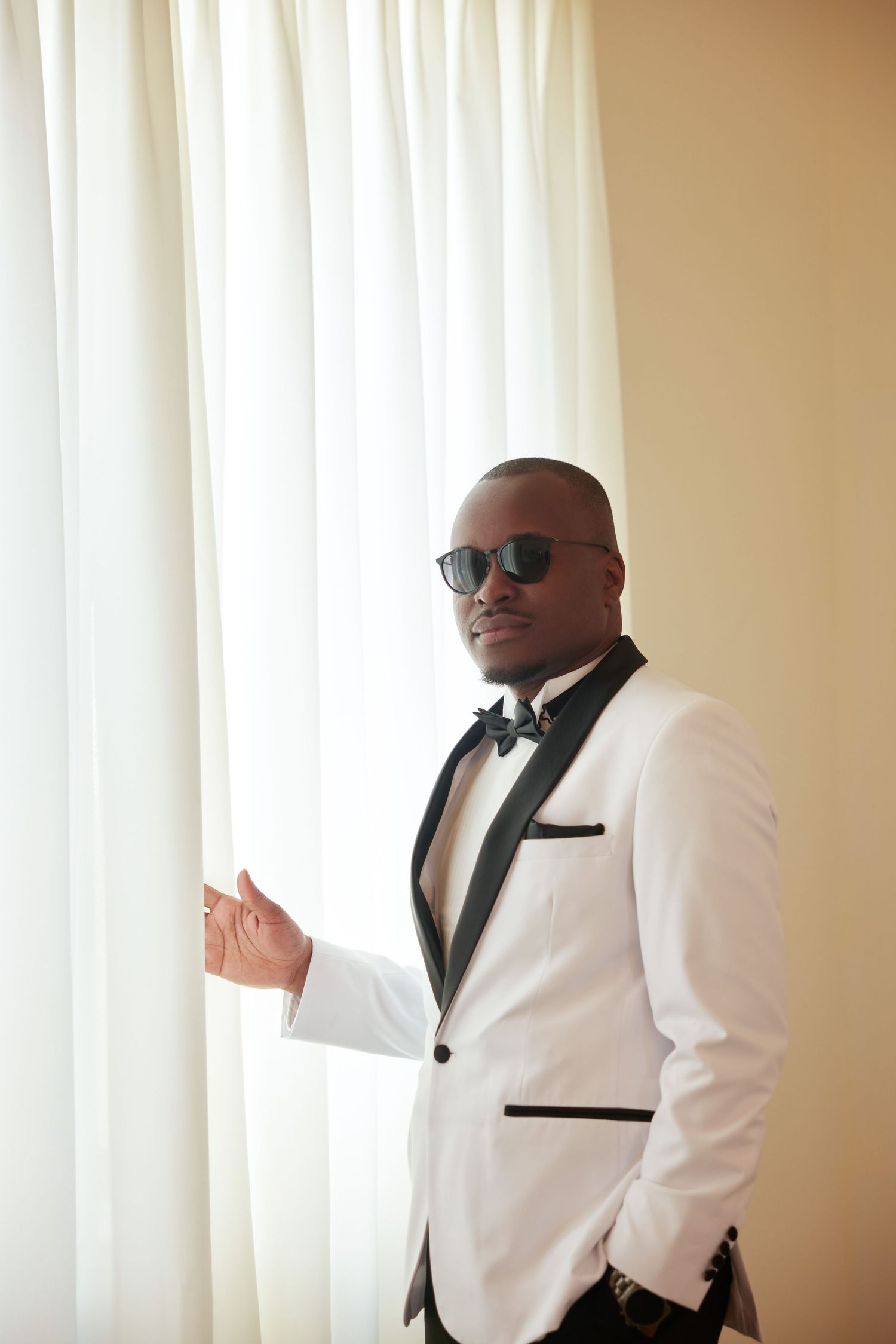 Stylish bald man in white tuxedo and sunglasses posing by white curtains indoors.