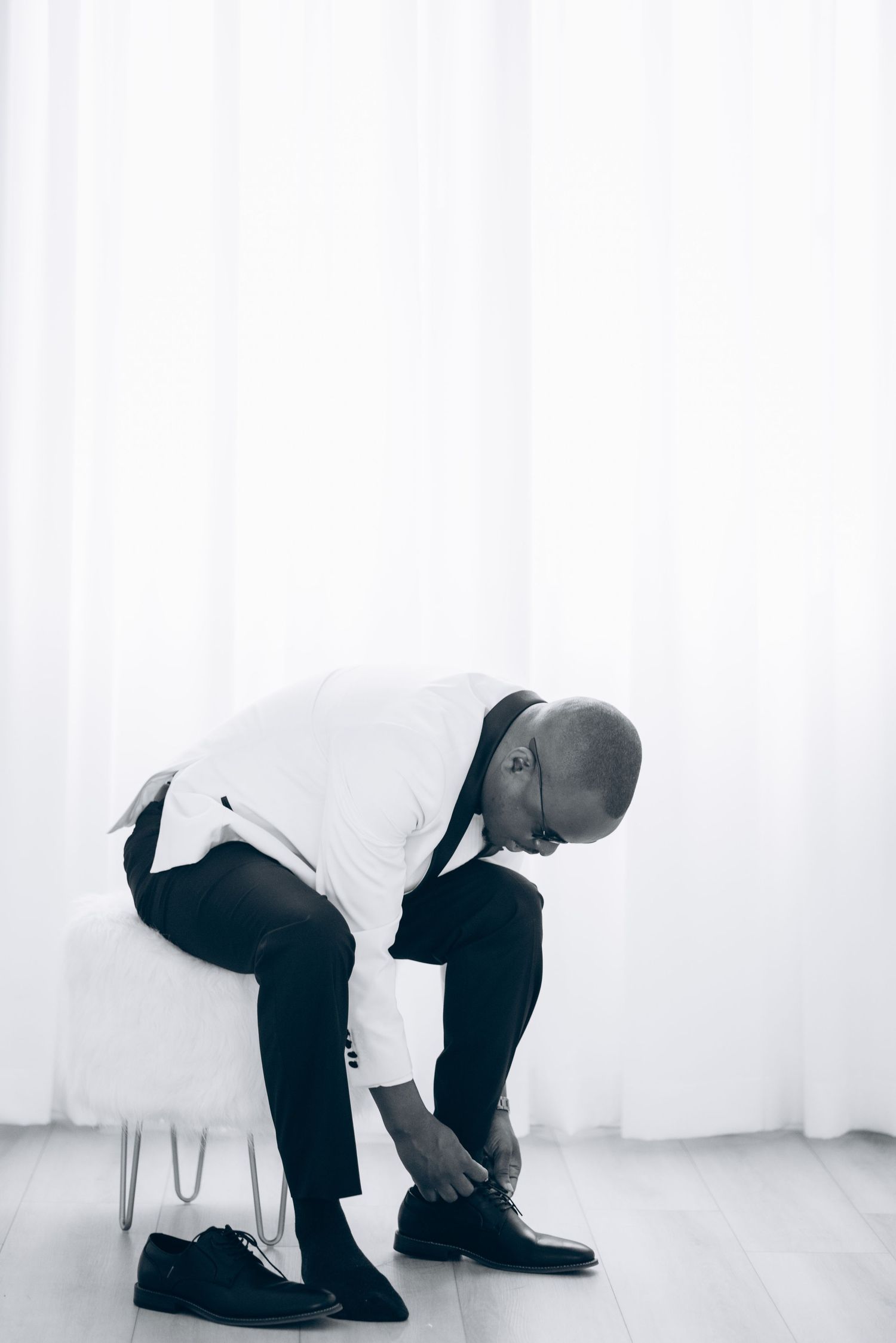 Black and white photo of a bald man in formal attire sitting on a stool tying his dress shoes.