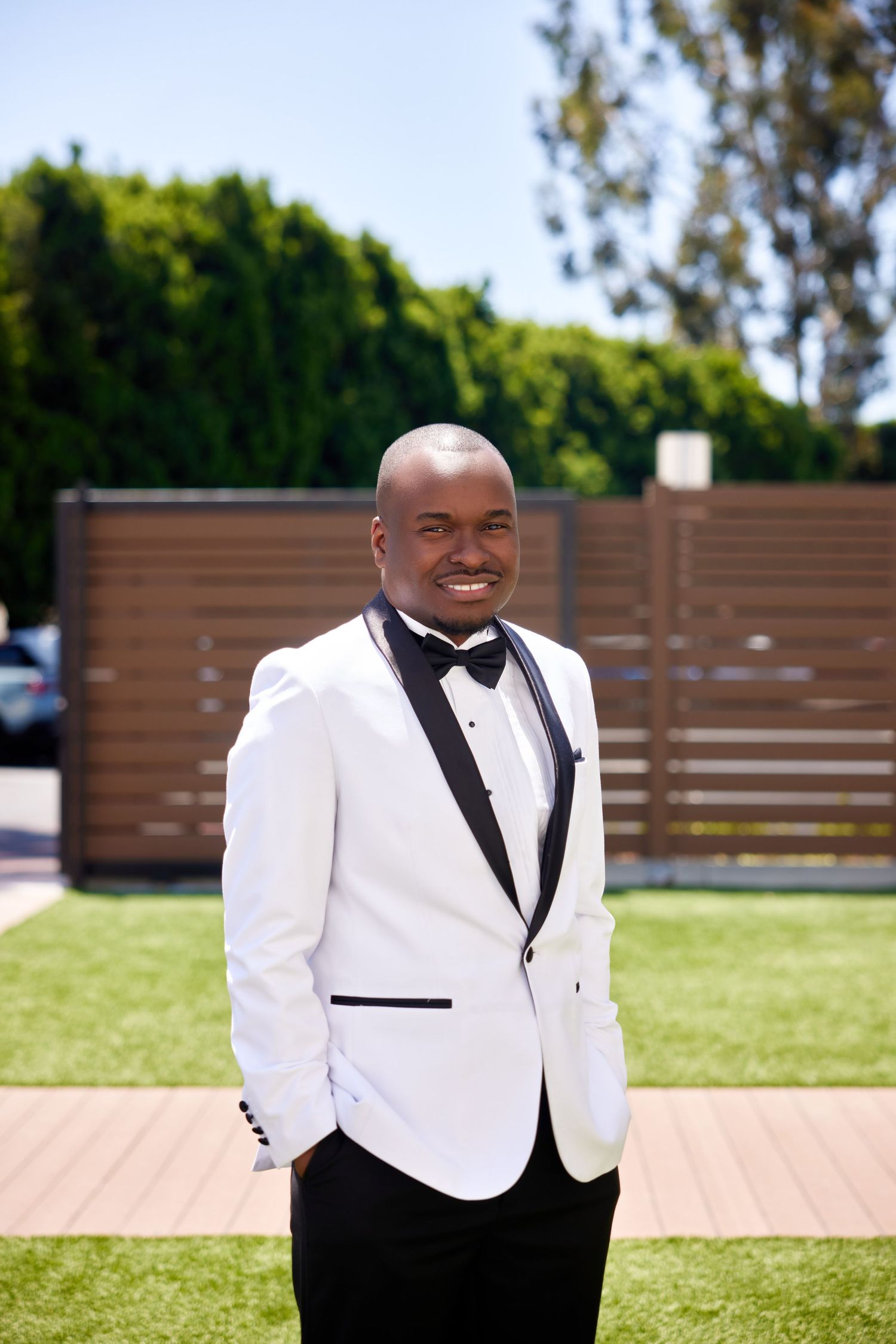 Smiling bald man in white tuxedo with black lapels and bow tie standing outdoors on green lawn.