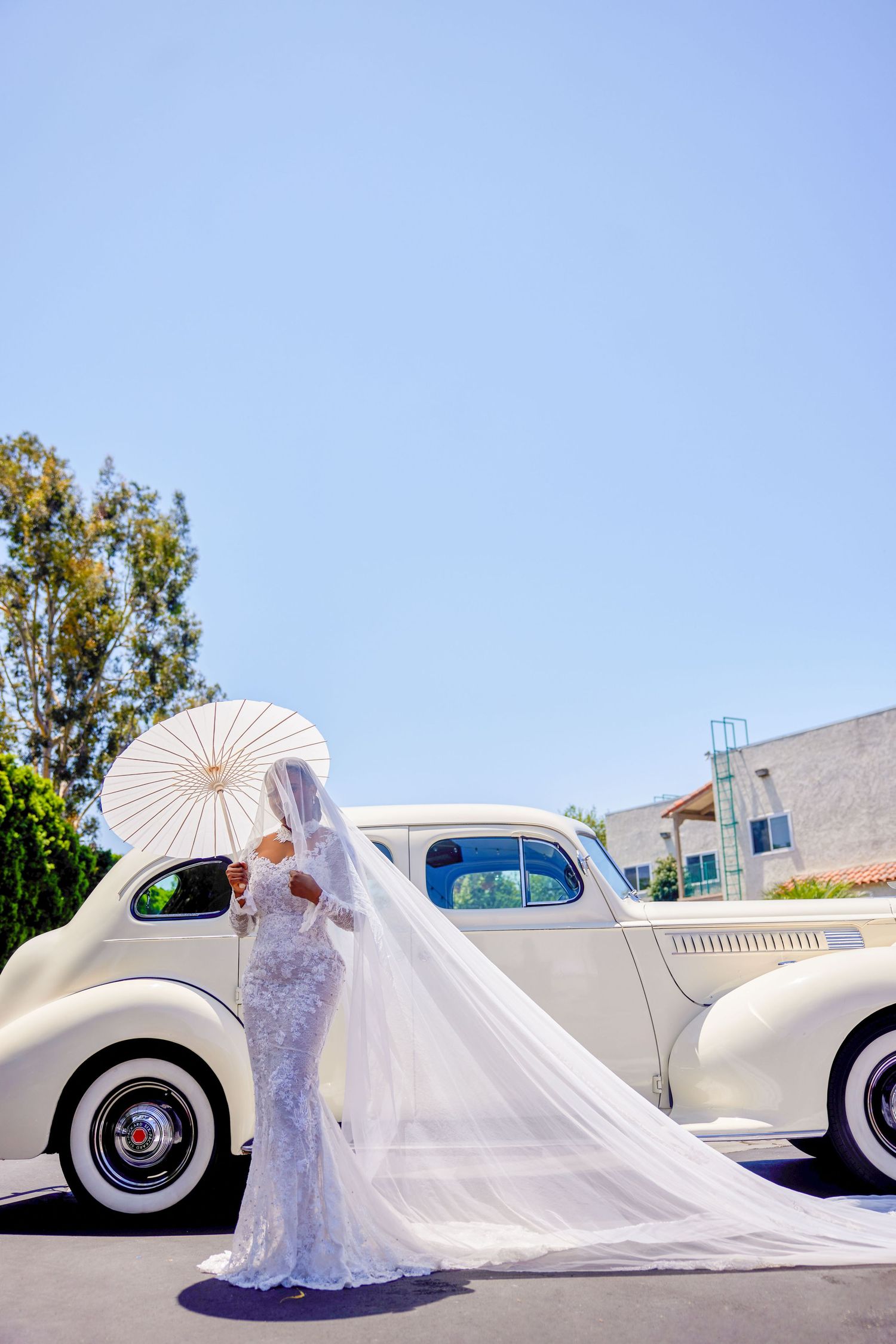 Bride in lace wedding gown with long veil holds white parasol beside vintage cream car on sunny day.