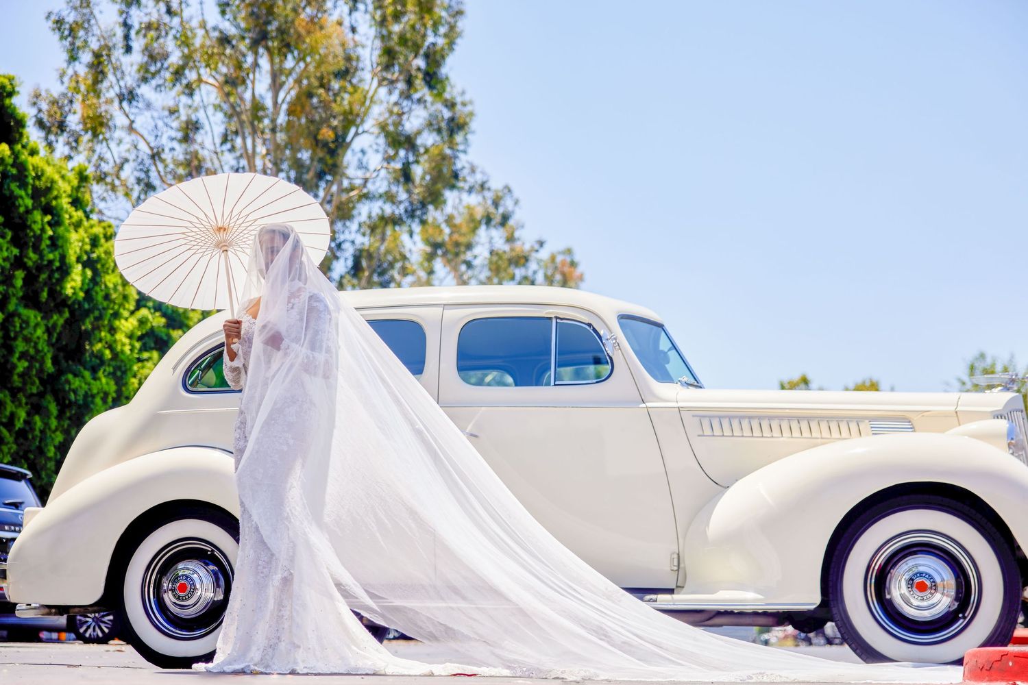 Bride in white gown holding parasol stands beside vintage cream classic car on sunny day.