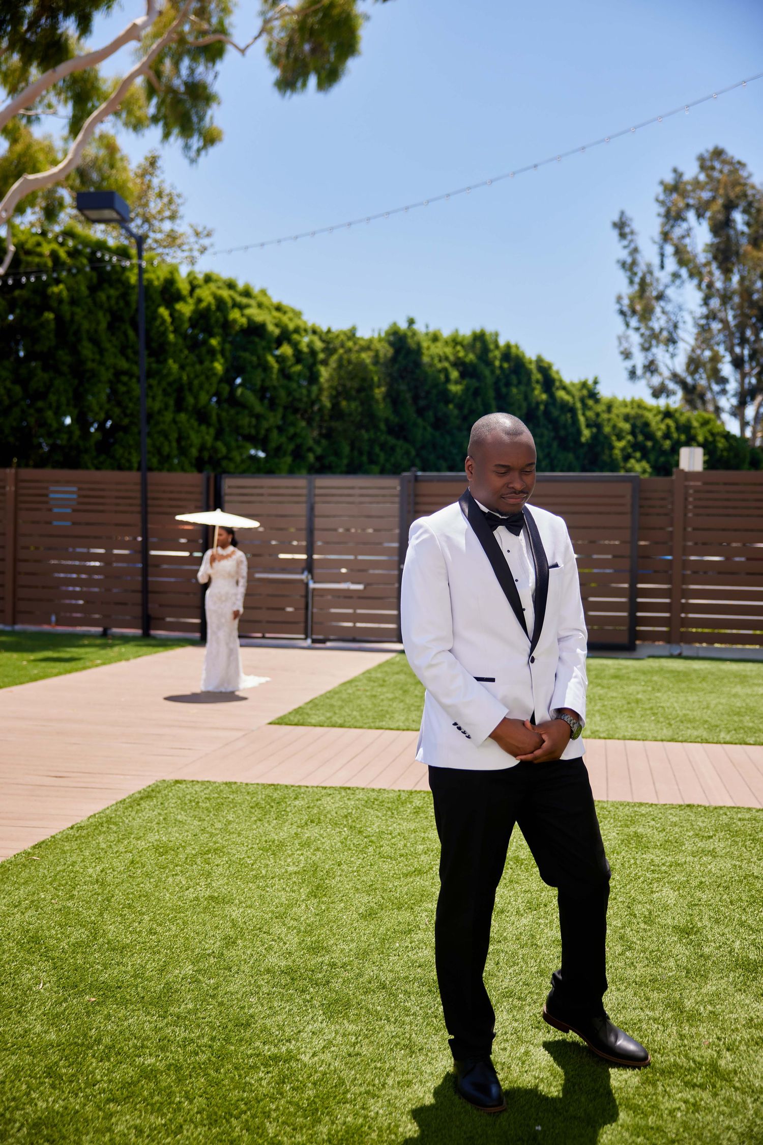Groom in white tuxedo stands on lawn as bride in white dress holds parasol in background at outdoor wedding.