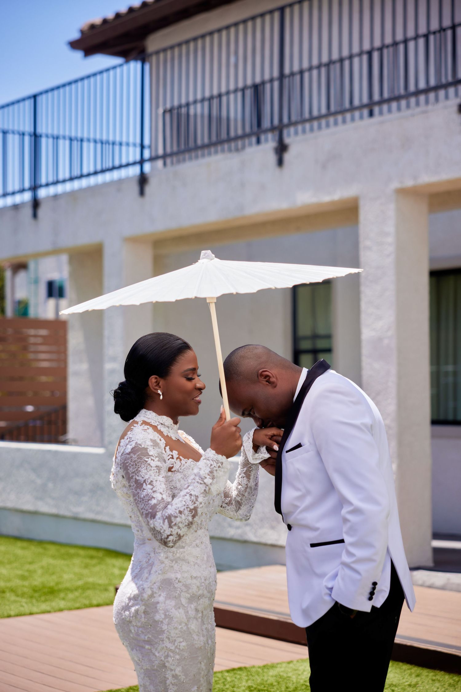 Black bride and groom share romantic moment under white parasol umbrella outside elegant venue on wedding day.