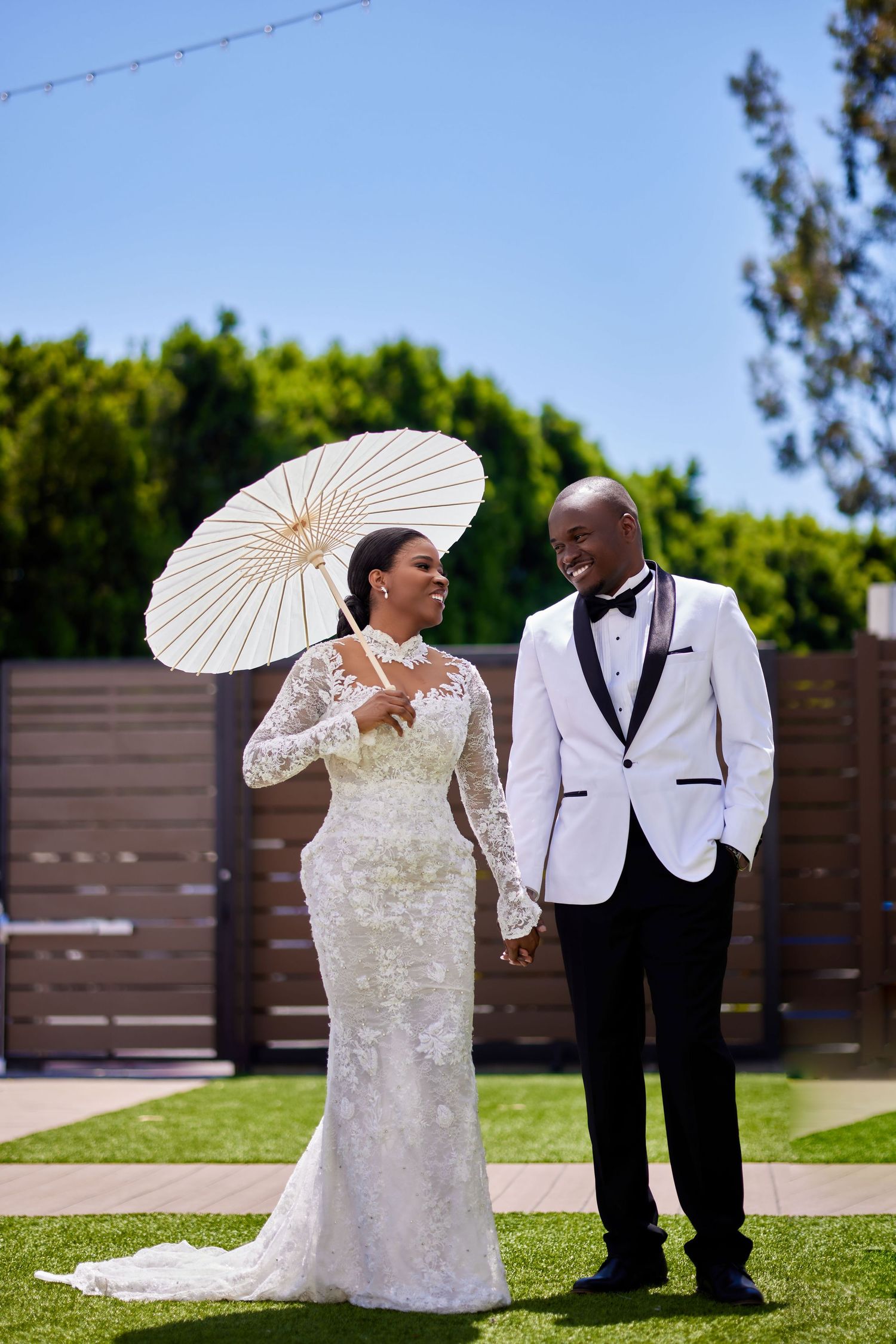 Smiling bride and groom stand on green lawn, bride holds white parasol wearing lace gown, groom in white tuxedo.