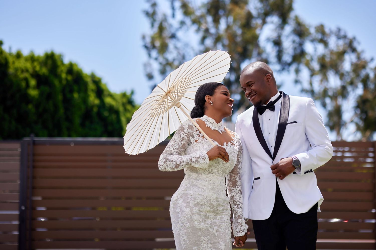 Elegant Black couple in wedding attire, bride holding white parasol, groom in white tuxedo, sharing a joyful moment outdoors.