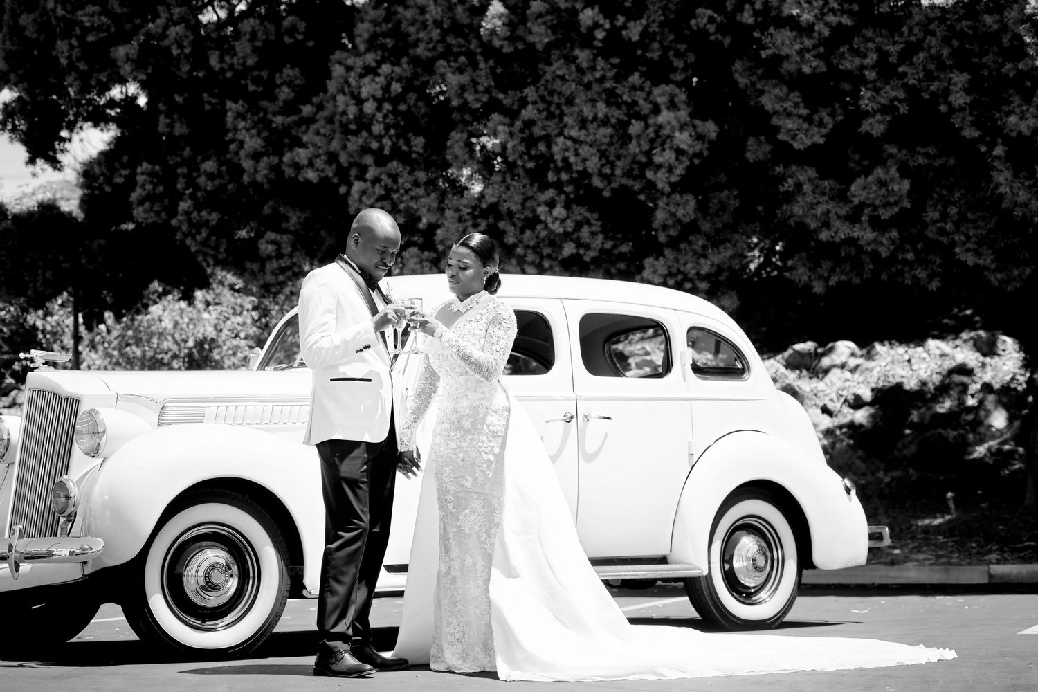 Black and white photo of bride and groom posing beside a vintage white classic car outdoors.