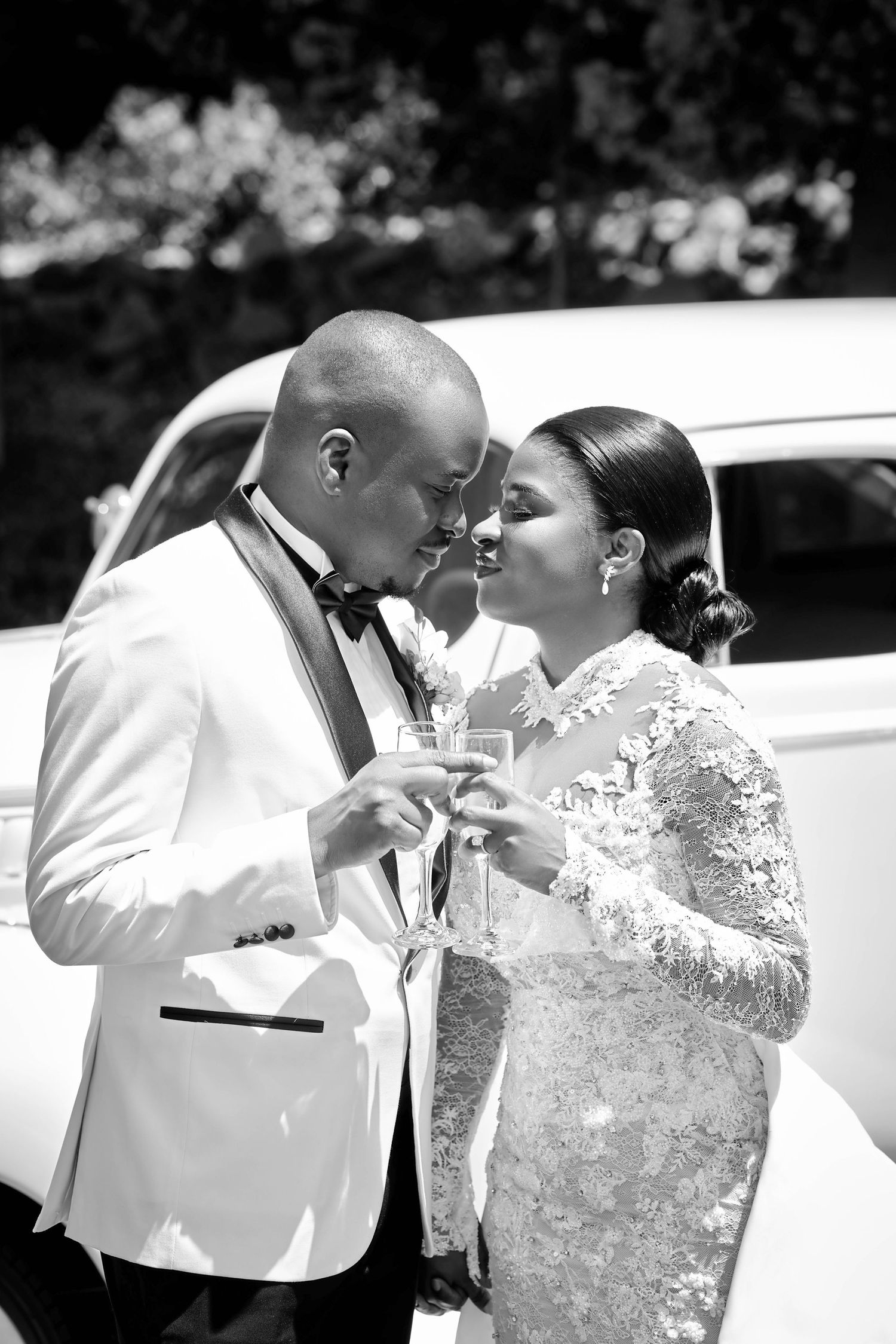 Bride and groom toasting with champagne glasses, foreheads touching, beside a vintage white car.