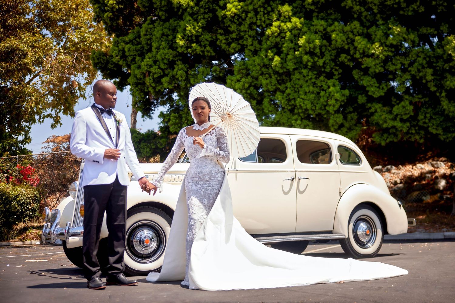 Bride and groom pose by a vintage cream car, bride in lace gown with parasol, groom in white tuxedo.