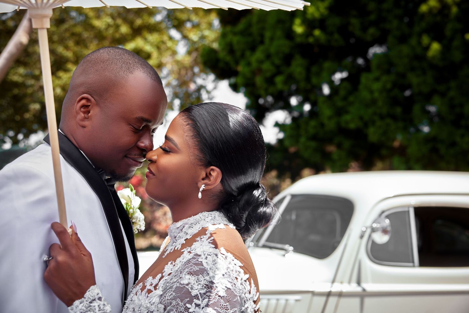A bride and groom share a romantic moment under a white umbrella beside a vintage car on their wedding day.