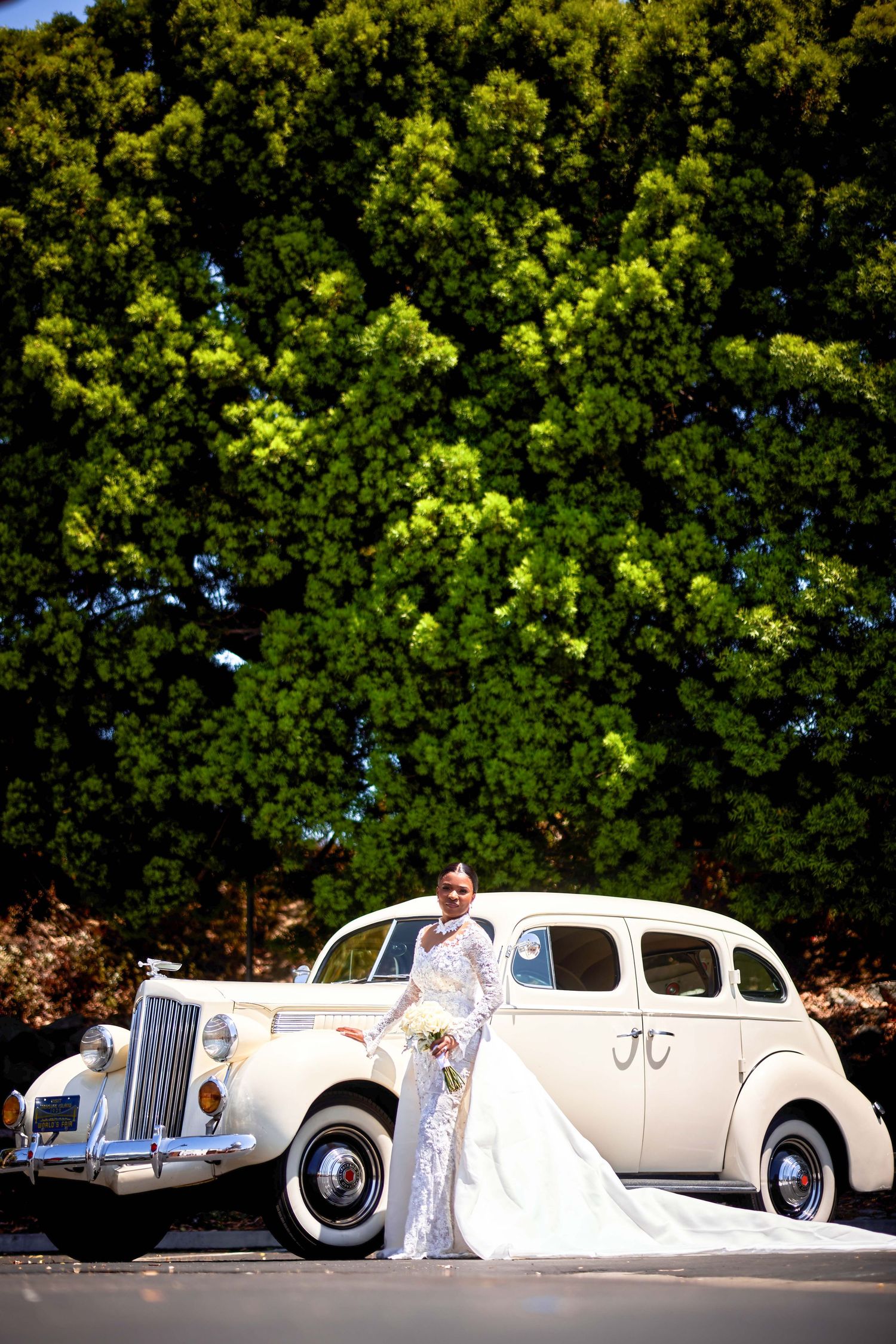 Bride in lace wedding dress posing beside vintage white classic car with lush green trees backdrop.
