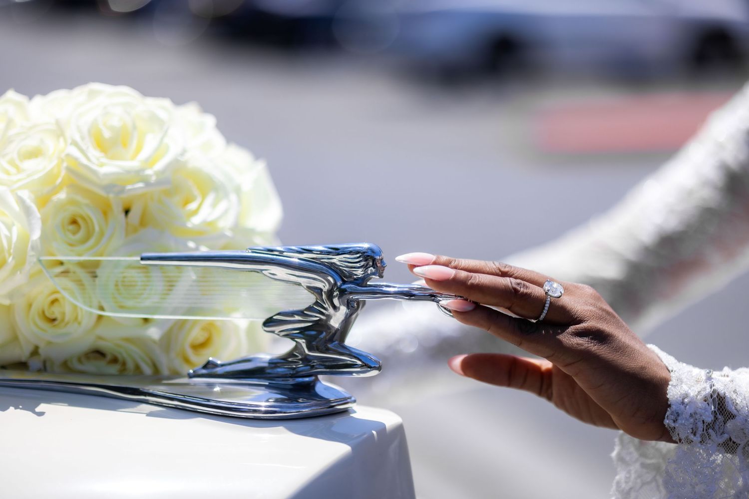 Bride's hand with wedding ring touching chrome hood ornament on white car beside bouquet of white roses.