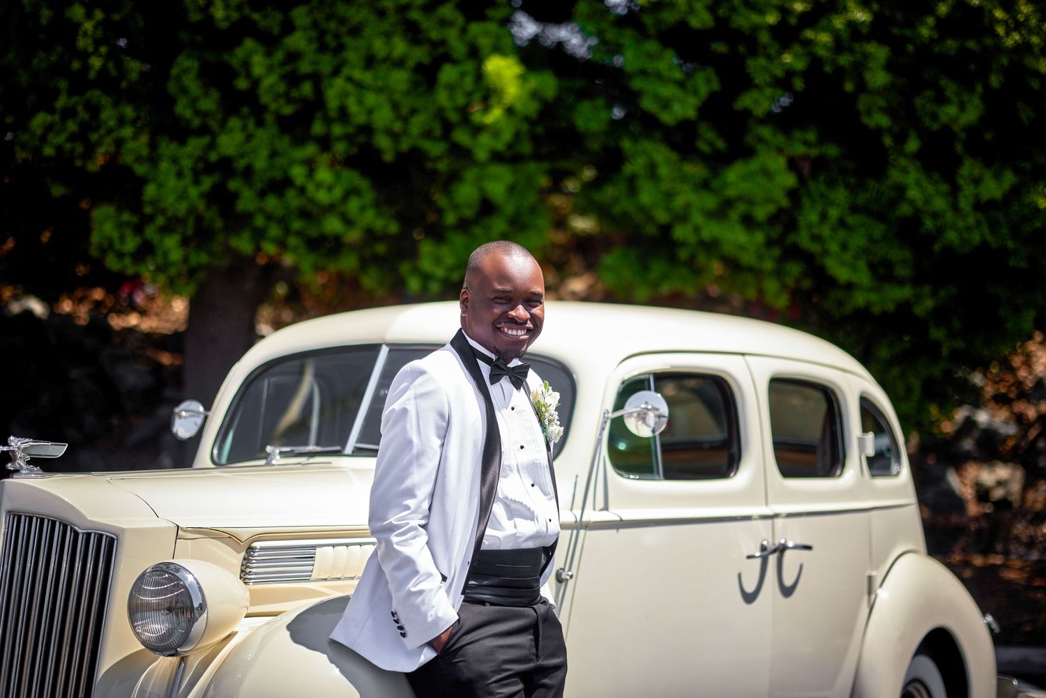 Smiling groom in white tuxedo leans against a classic cream vintage car with floral decor and monogram detail.