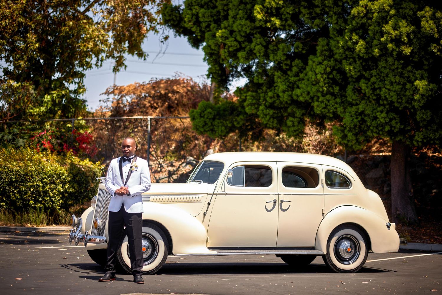 Groom in white tuxedo poses beside a classic cream vintage car decorated with initials in a sunny outdoor setting.
