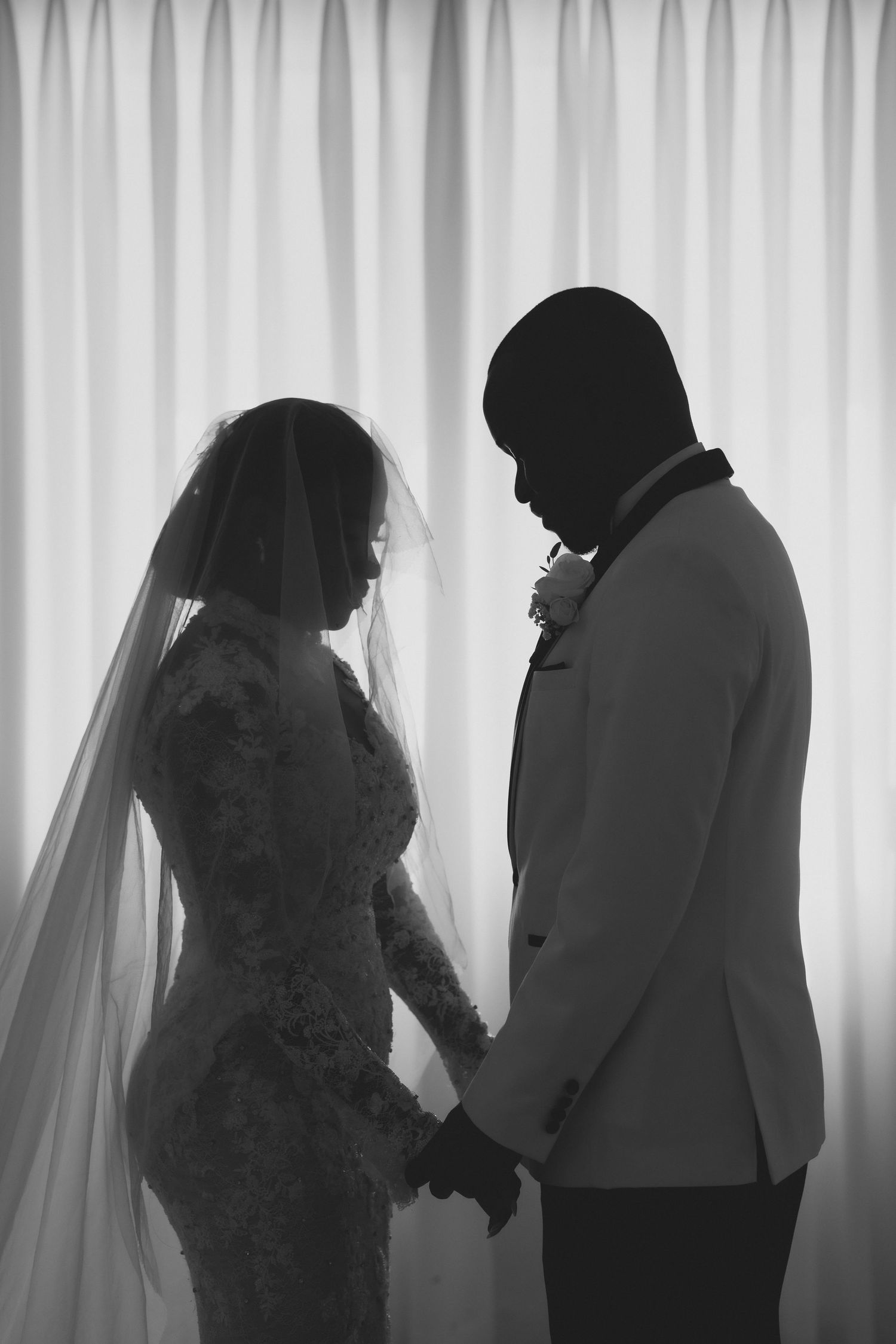 Black and white silhouette of bride in lace gown and veil holding hands with groom in suit before sheer curtains.