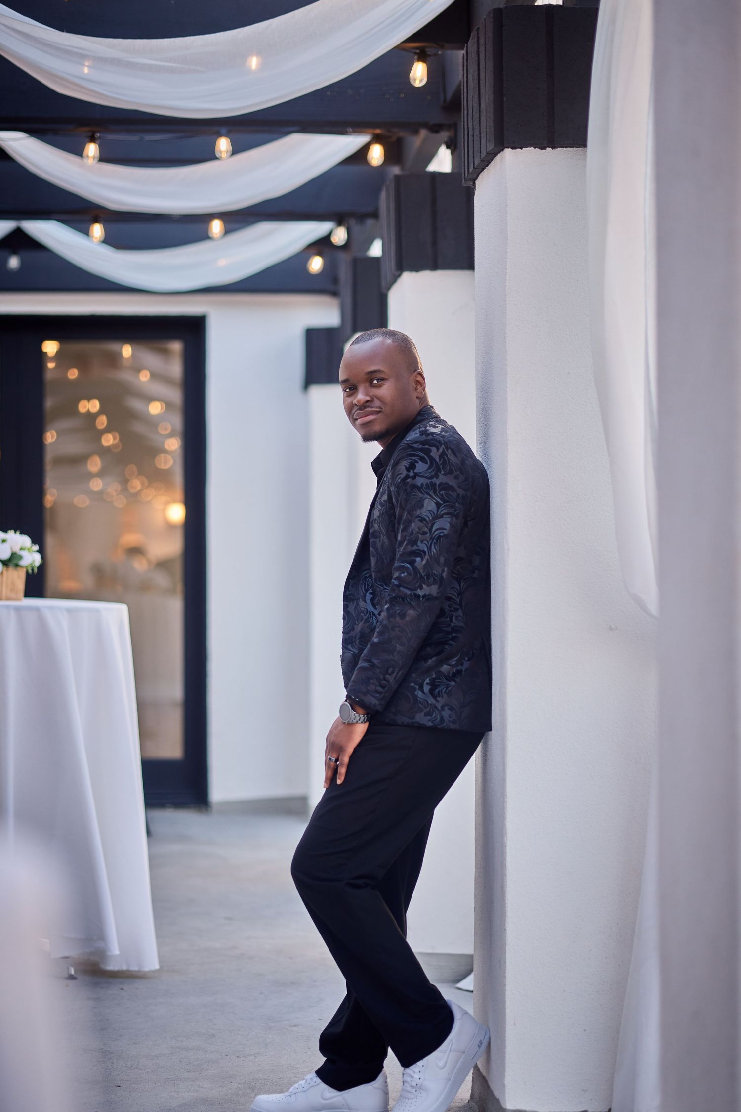 Stylish man in floral blazer and black pants leaning against a white pillar in an elegant event venue.