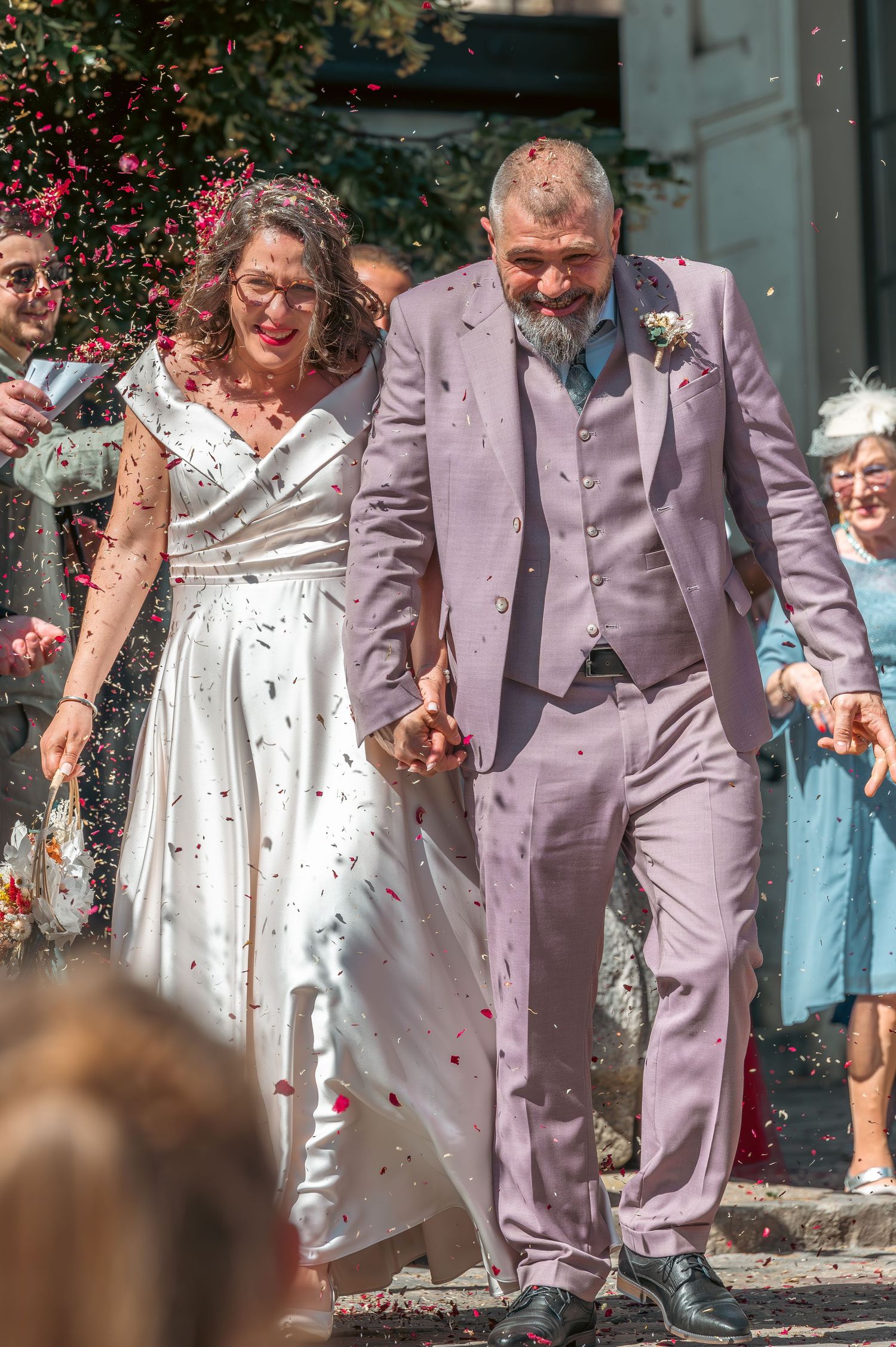 La mariée et le marié marchent main dans la main dans la douche de confettis à l'extérieur du lieu du mariage par une journée ensoleillée.