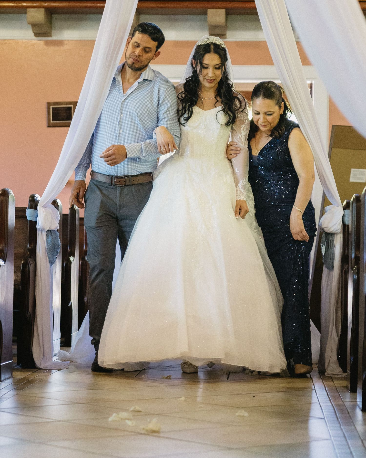 Bride in white ball gown walks down church aisle escorted by man in blue shirt and woman in navy sequin dress.