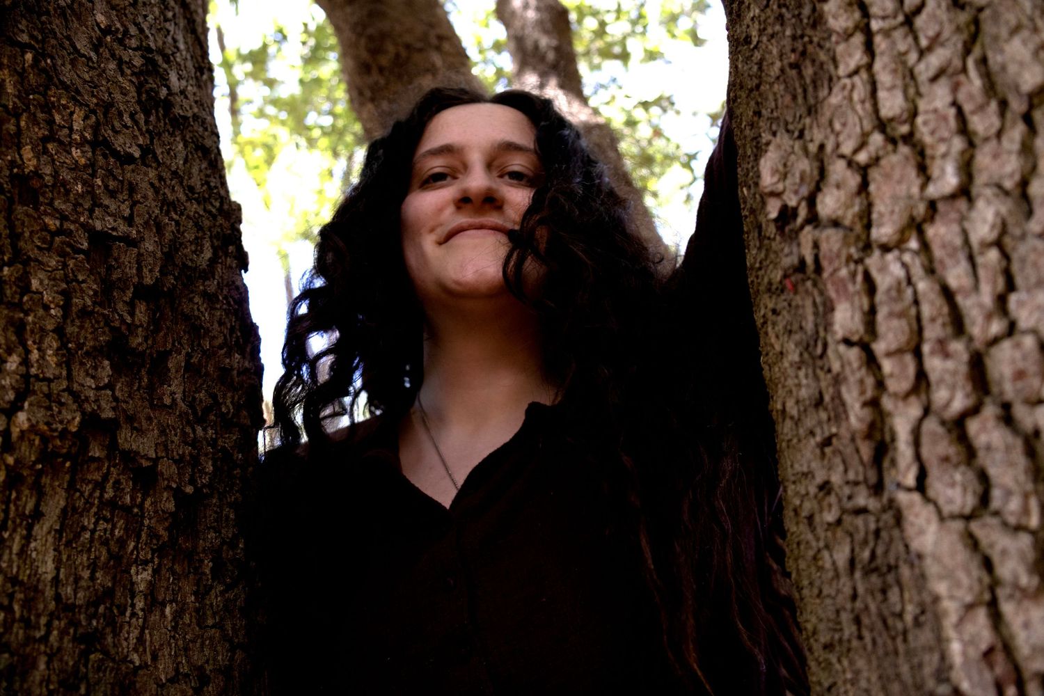 Young woman with curly dark hair smiling peacefully between two tree trunks in a sunlit forest.