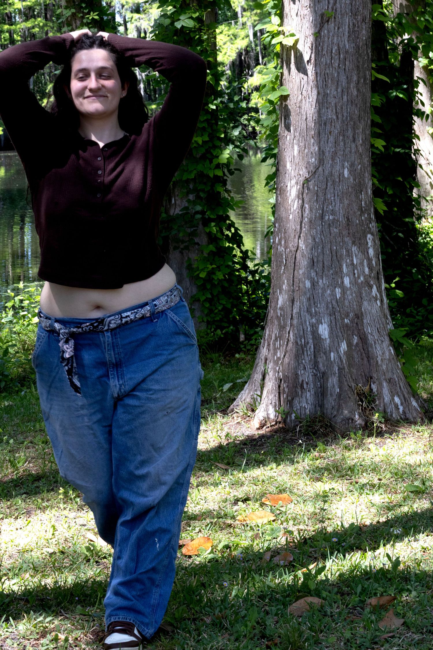 Smiling young woman in dark top and baggy jeans poses outdoors in a sunny green park near a tree.