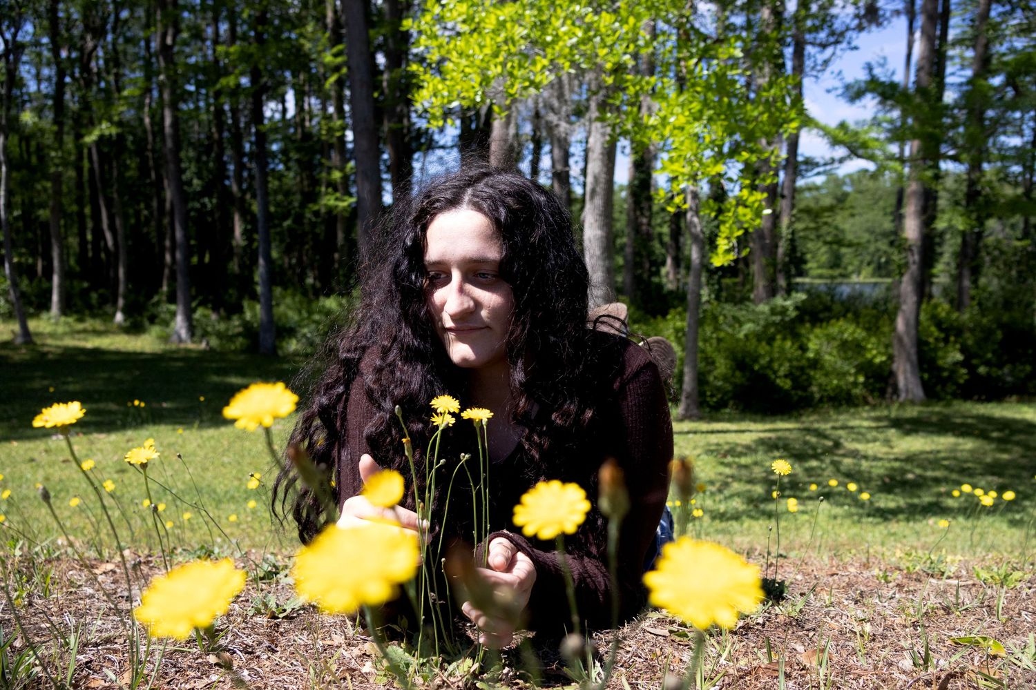 A dark-haired woman crouches among bright yellow dandelion flowers in a sunny green meadow.