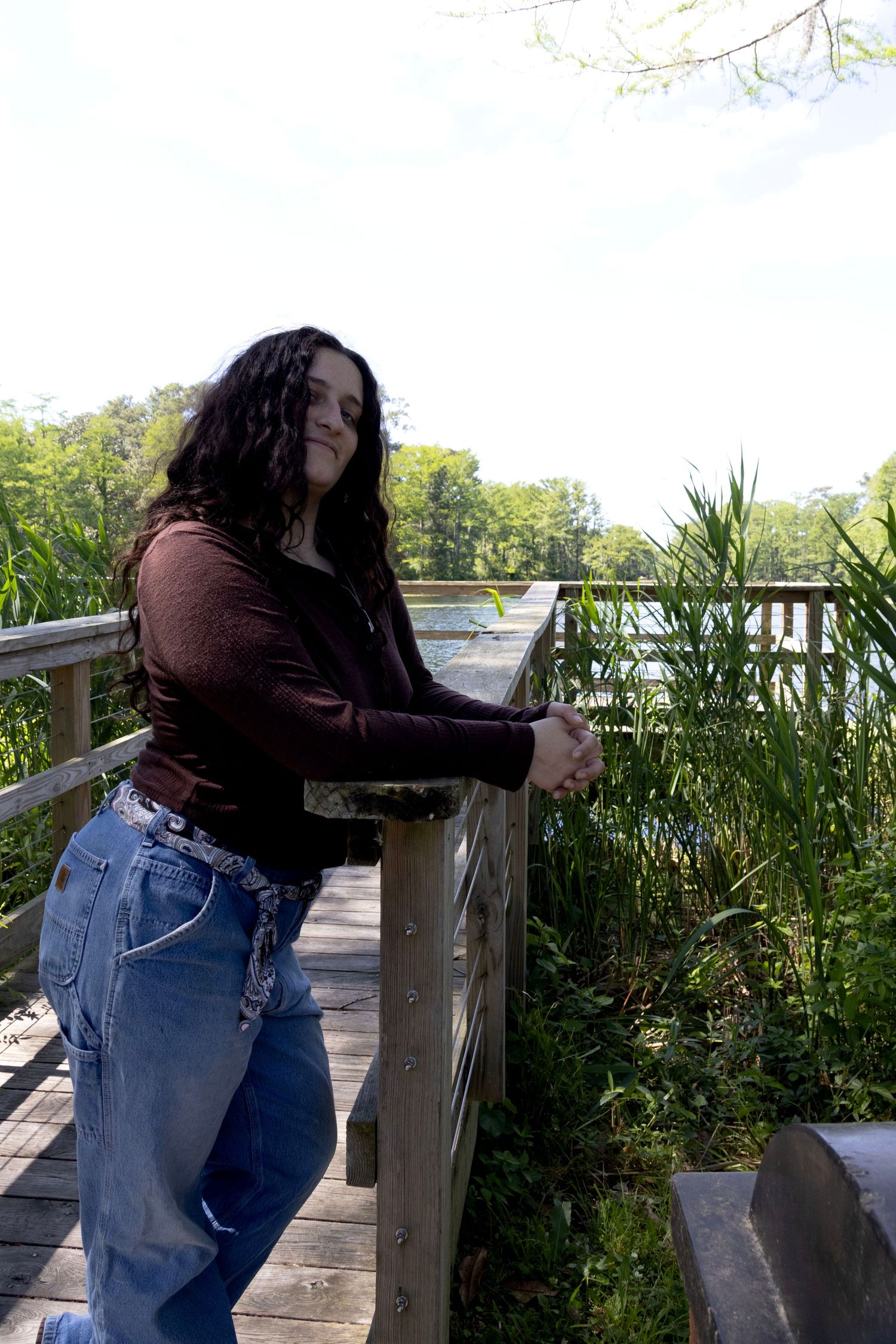 Young woman with dark curly hair in brown top and jeans leaning on wooden railing of nature boardwalk.