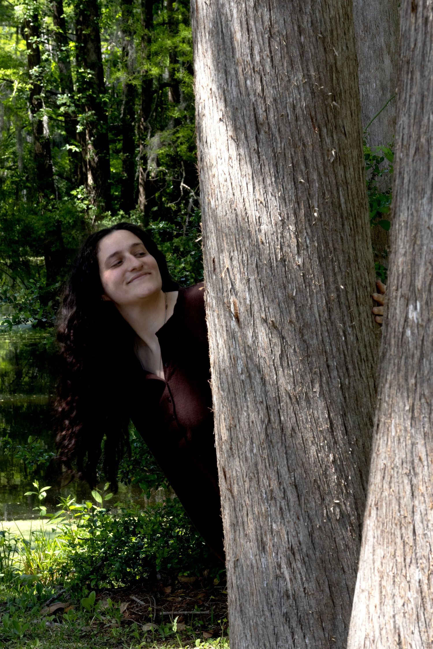 A smiling woman with long dark hair peeks out from behind a large tree trunk in a lush green forest.