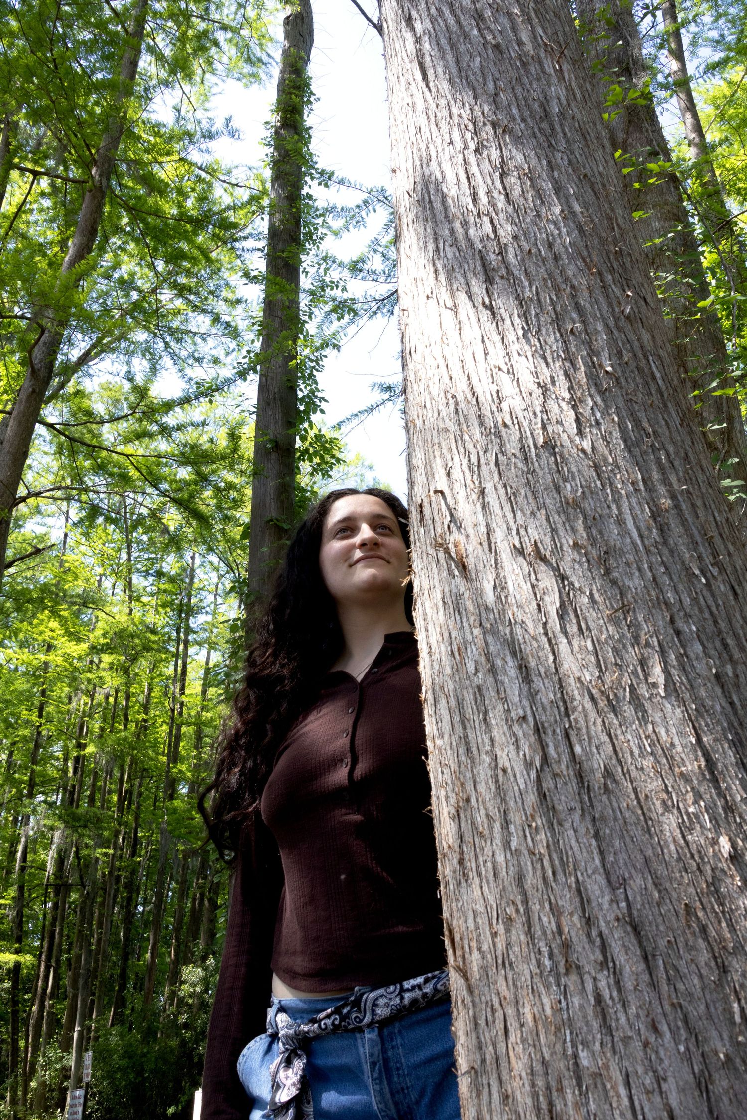 Young woman with long dark hair leans against a large tree trunk in a lush green forest.