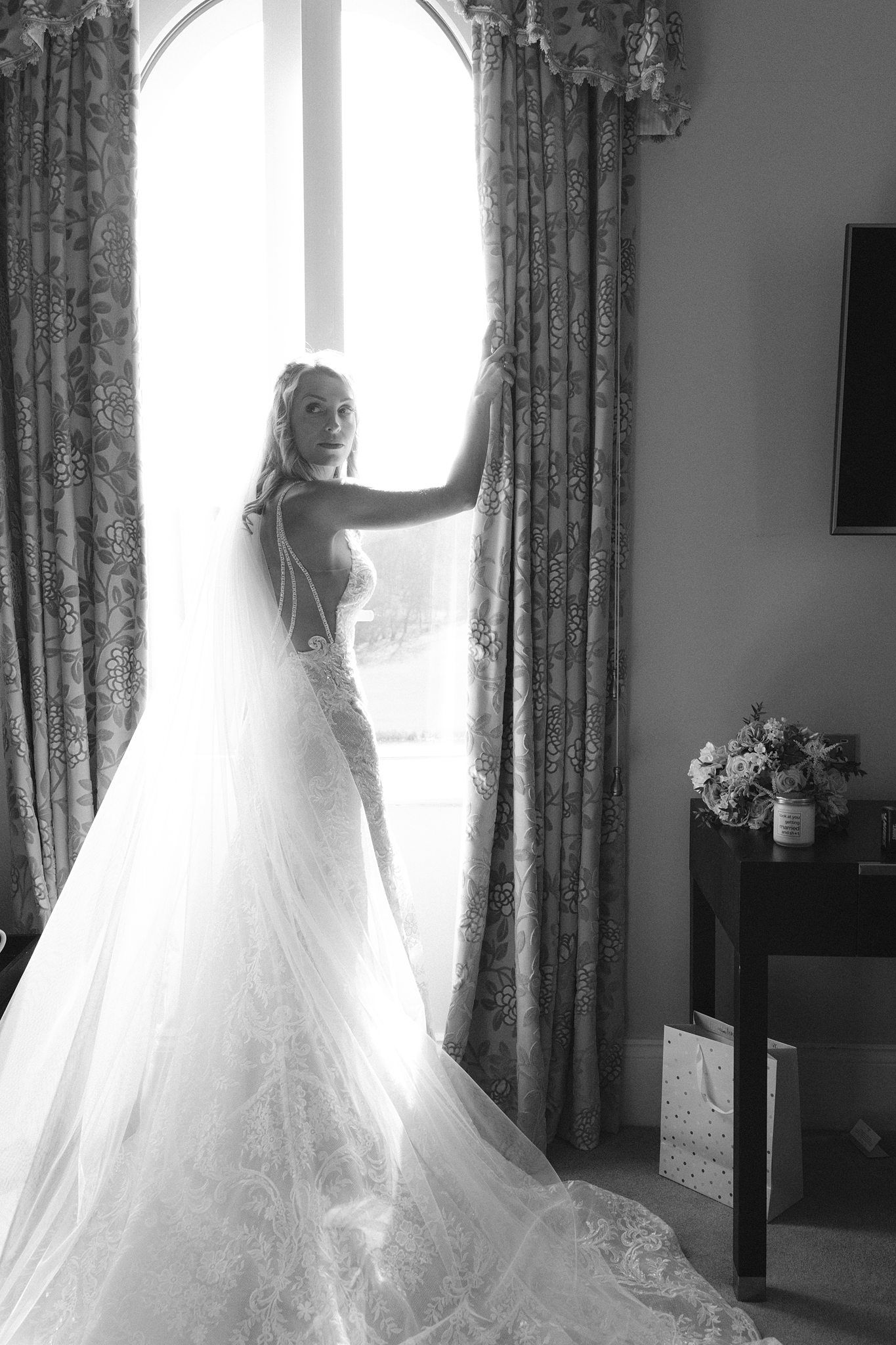 Bride in lace wedding gown with veil stands by window in black and white portrait photo.